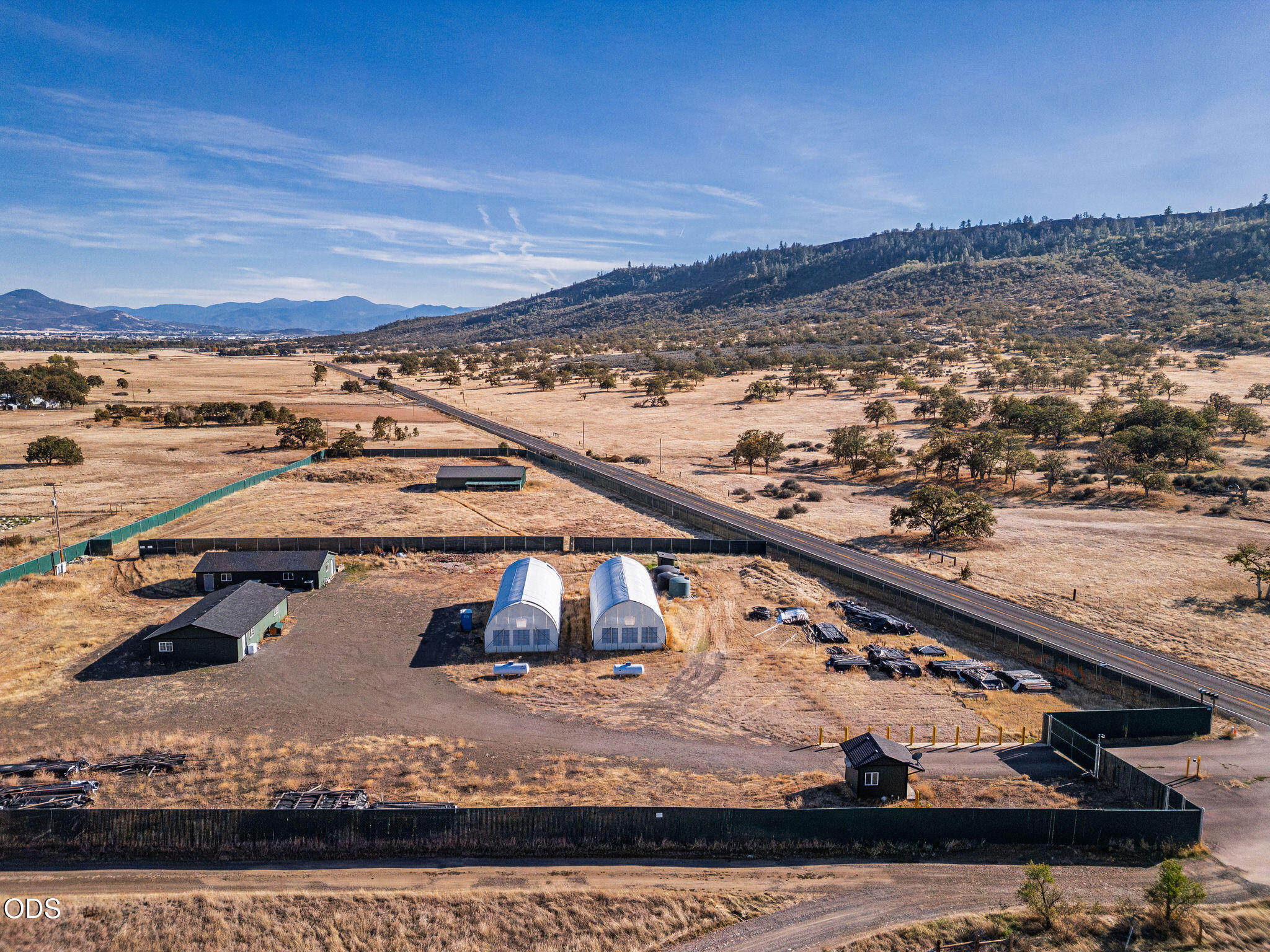 Aerial view of fenced rural property with two greenhouses, outbuildings, and mountain views.