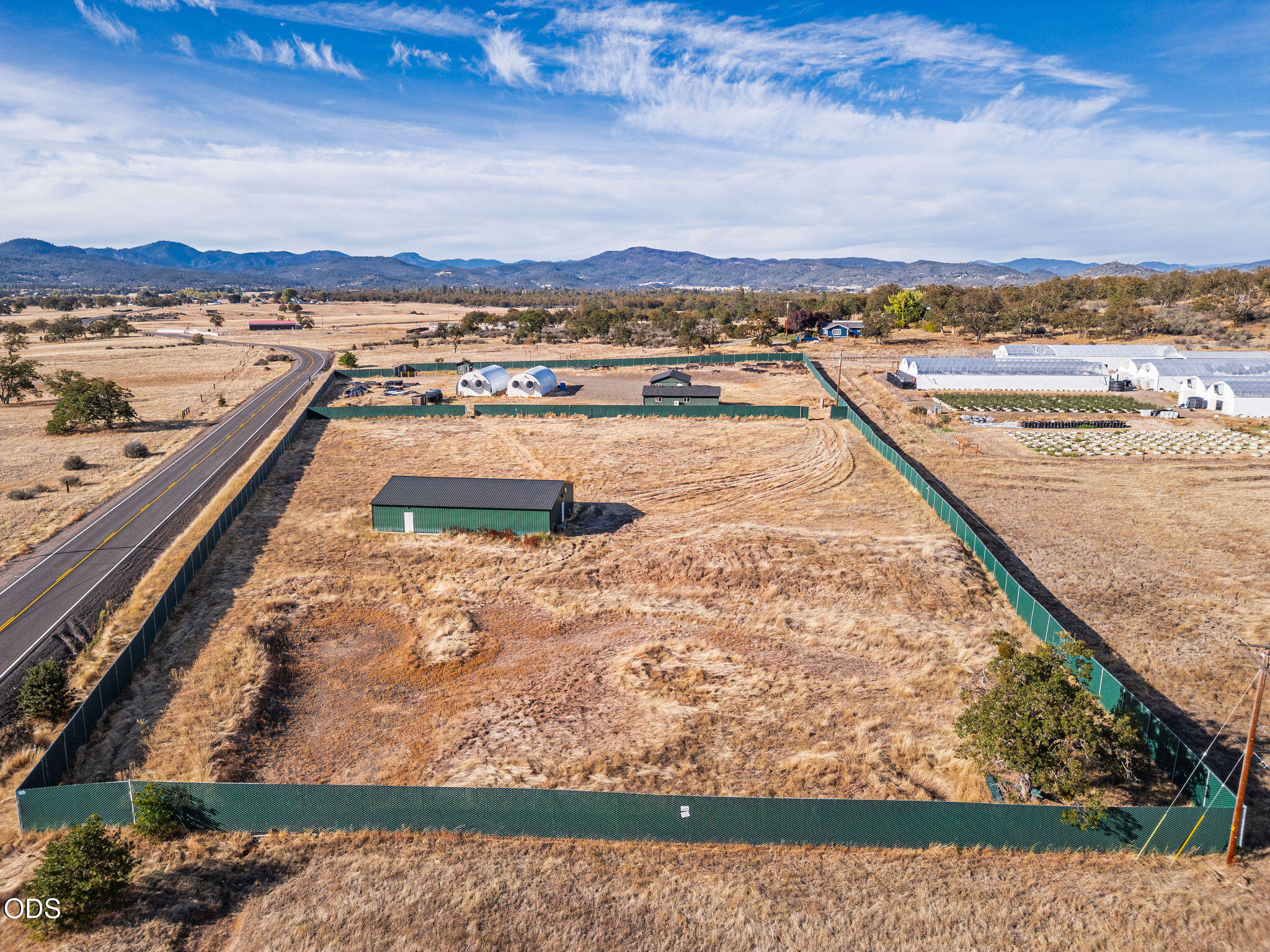 Fenced rural property with green outbuildings and greenhouses, beside a road with mountain ridges beyond.