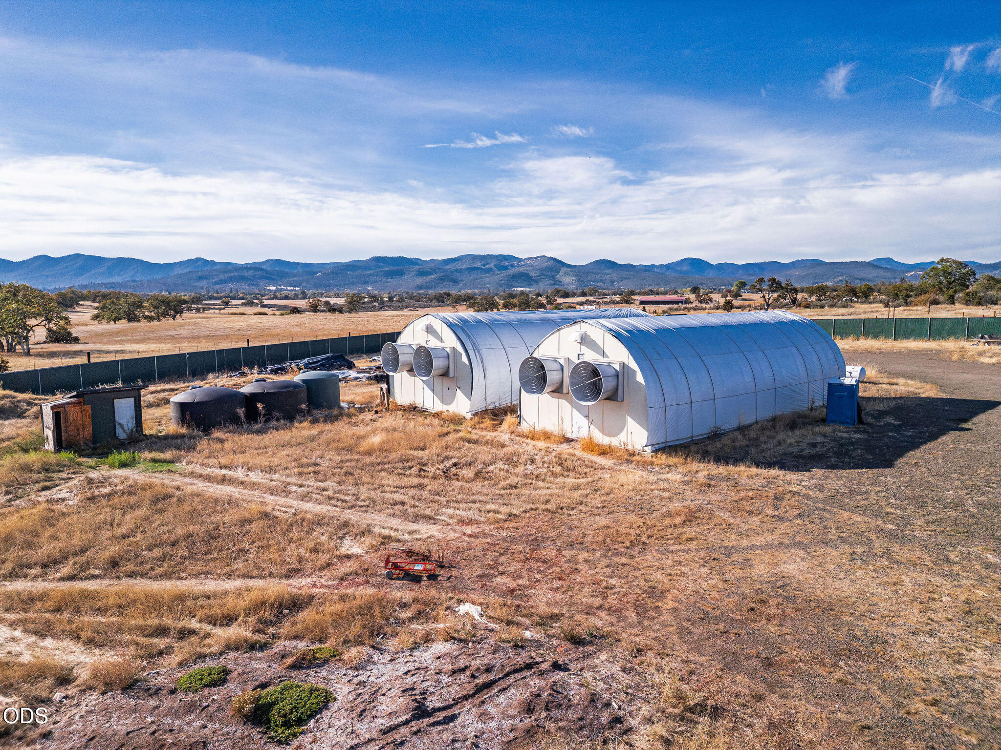 Two white agricultural buildings with large ventilation fans stand on dry land, with mountains in the distance.
