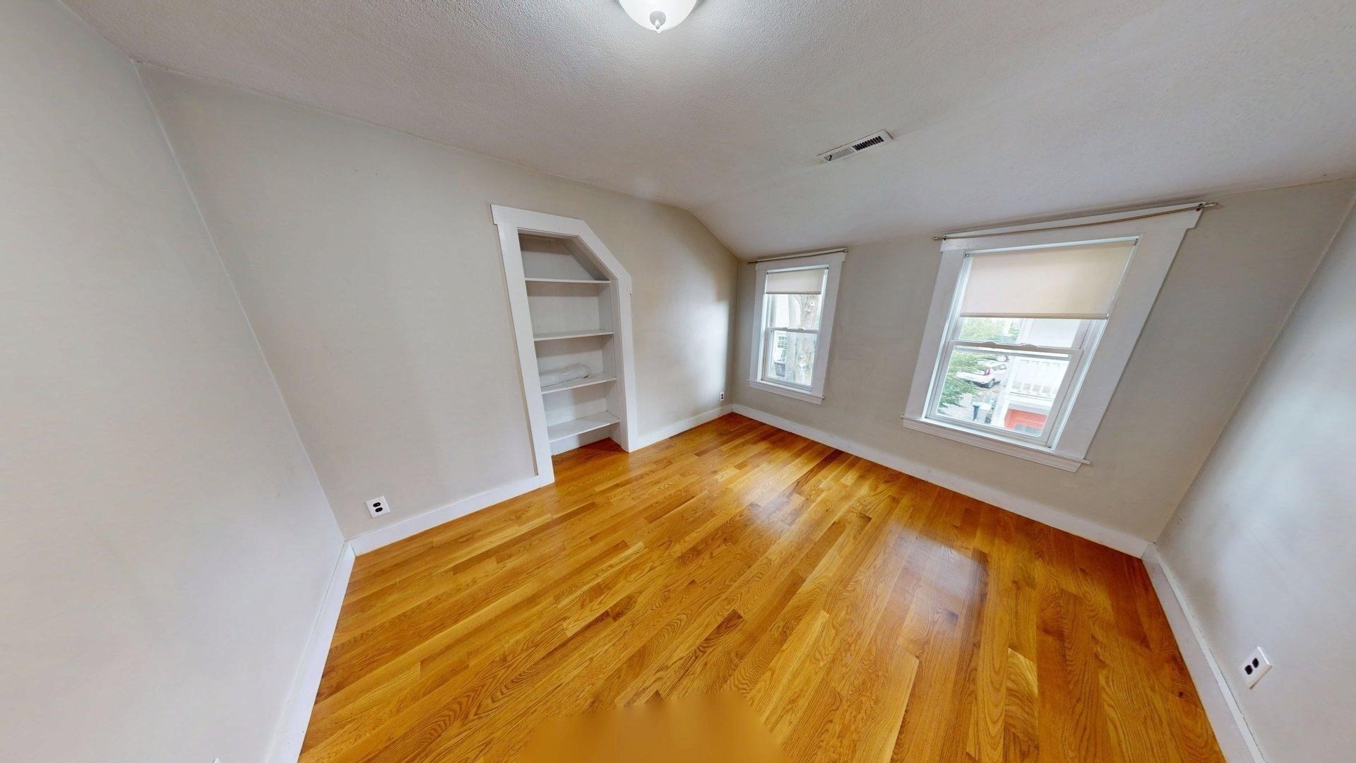 Empty room with sloped ceilings, hardwood floors, built-in shelves, and two shaded windows.
