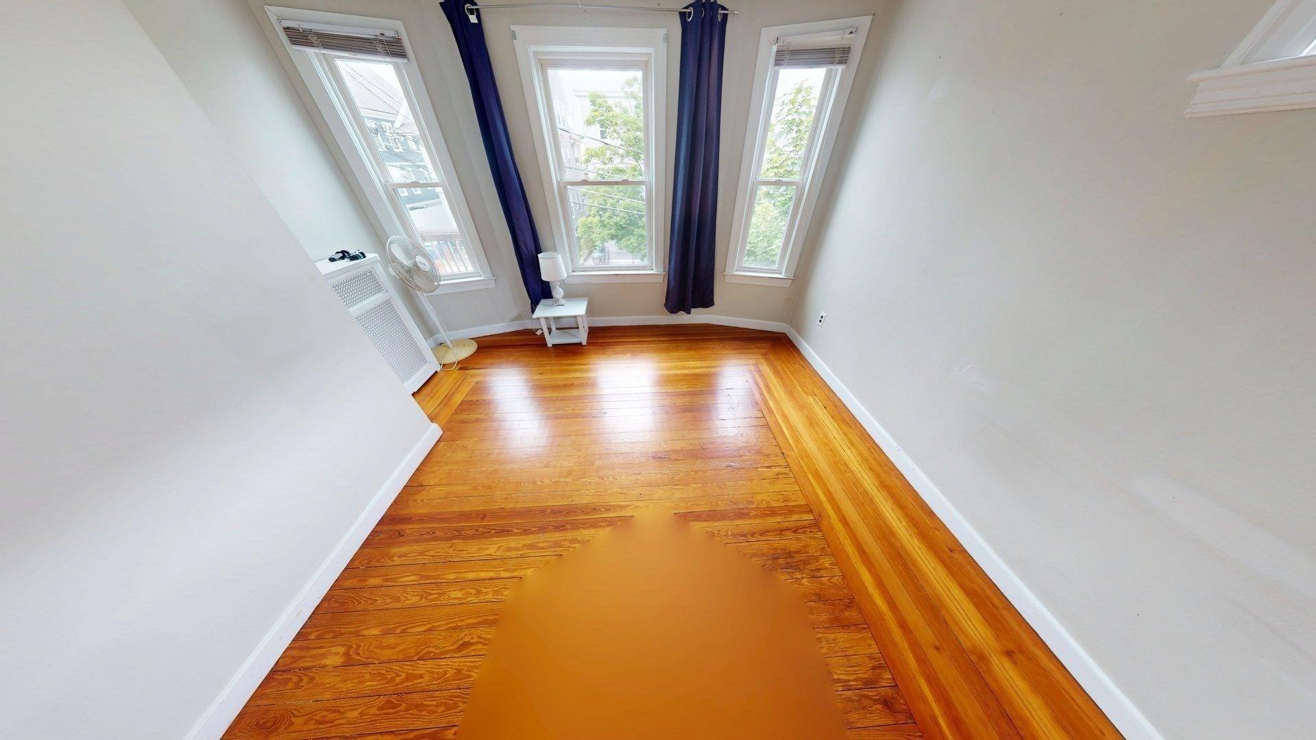 Bright room with polished hardwood floors, three tall windows, dark curtains, and a standing fan.