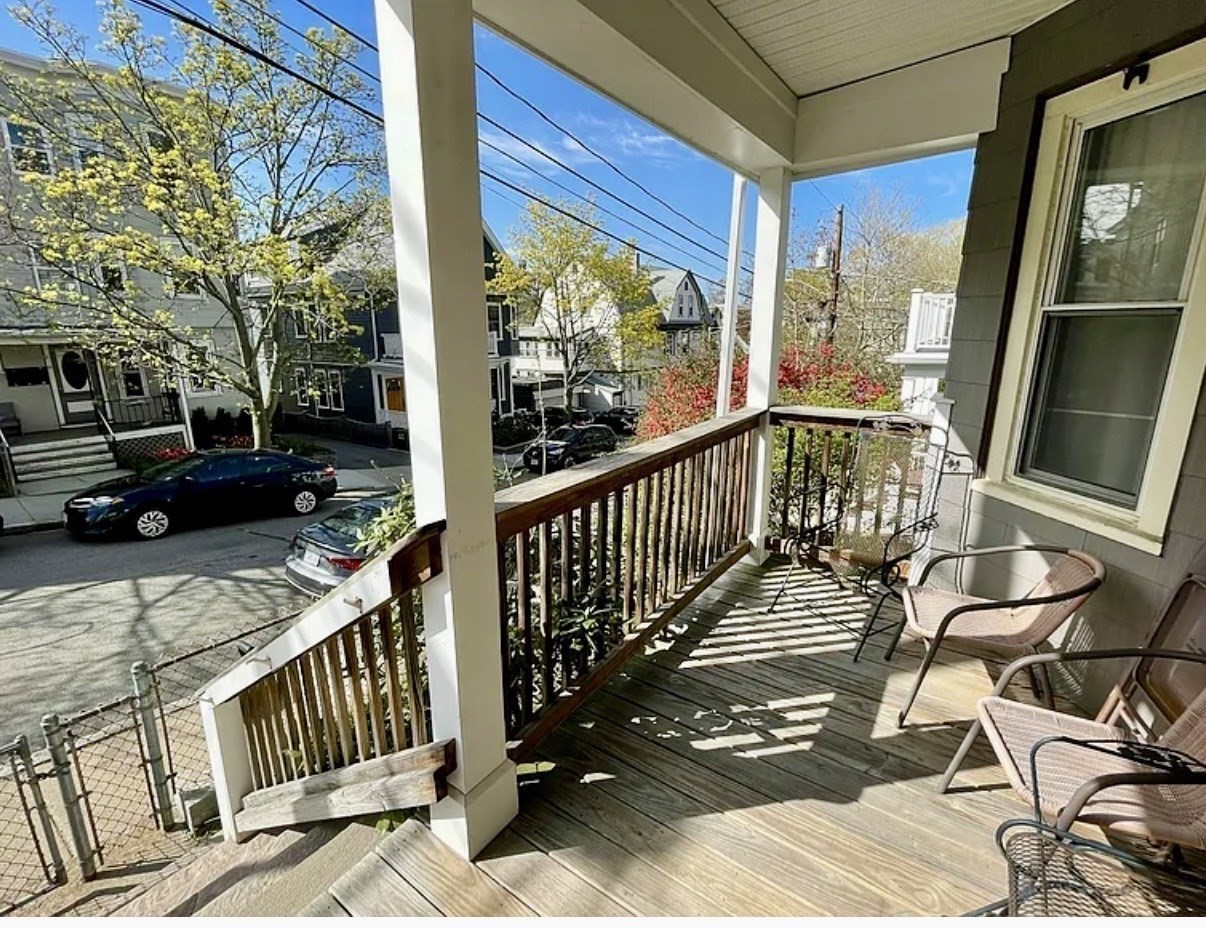 Covered wooden porch with chairs and steps overlooking a tree-lined residential street.
