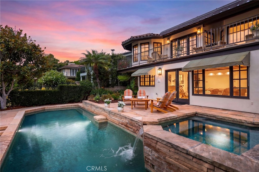 Two-story home beside a stone-edged pool and raised spa at dusk, with patio seating and a balcony.