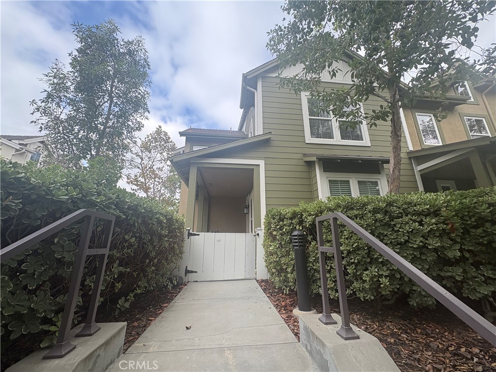 Two-story sage-green home with a gated walkway bordered by hedges and metal railings.