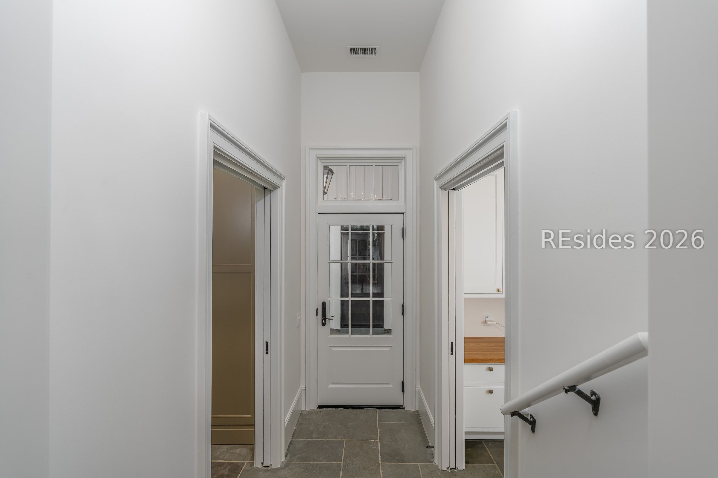 White hallway with gray tile flooring, a glass-paneled door, and a stair handrail.