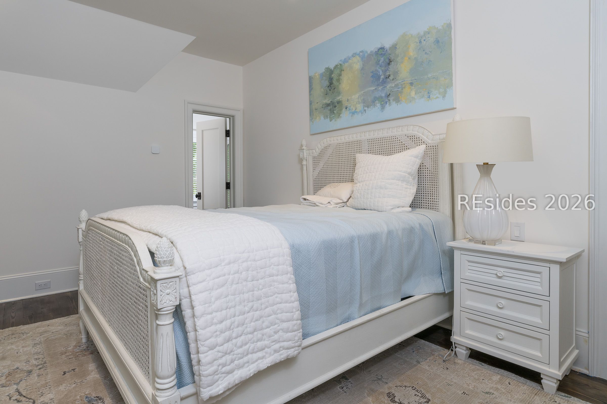 Bright bedroom with a white cane bed, pale blue bedding, abstract art, and white nightstand.