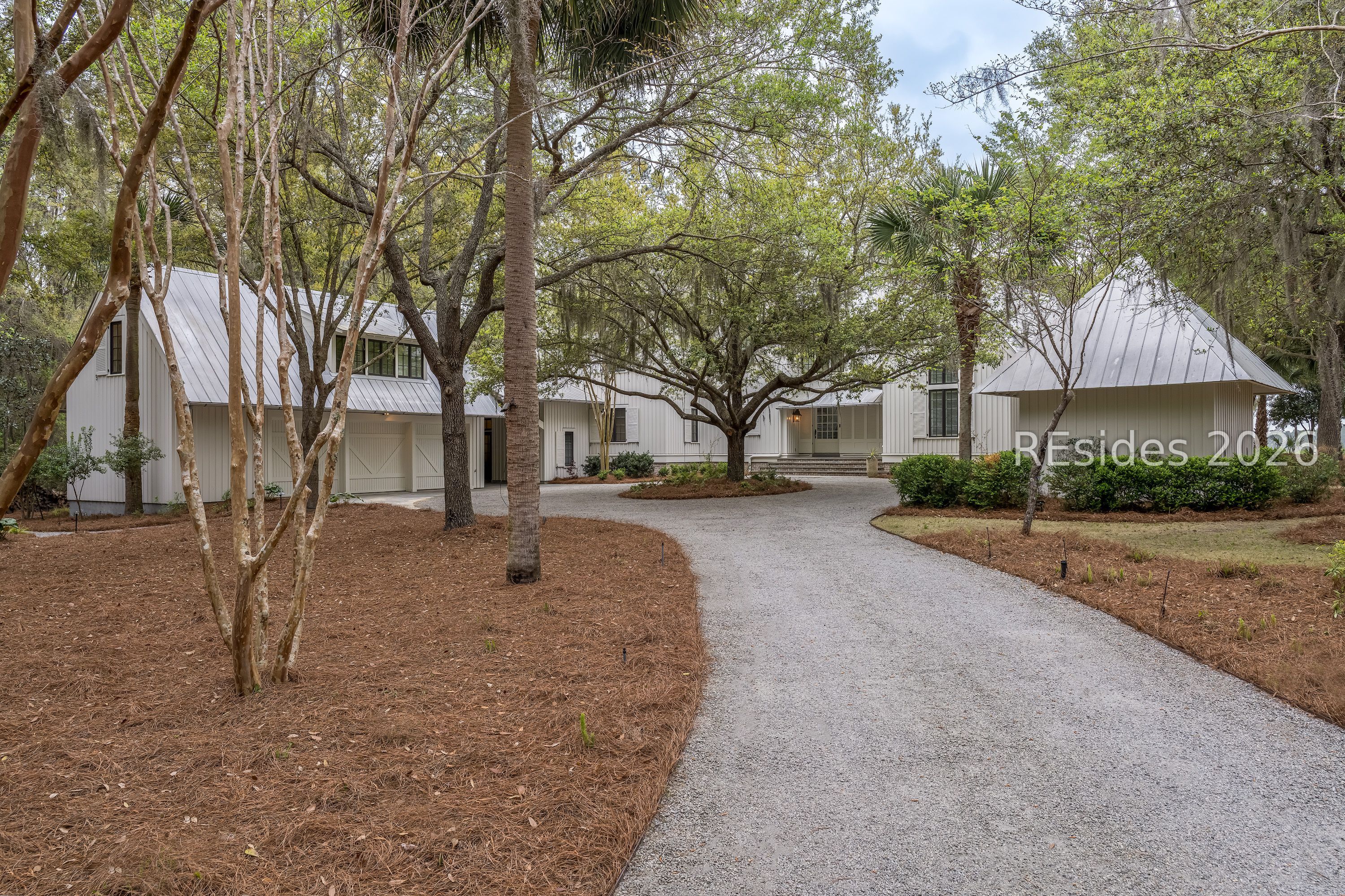 White home with metal roofs and a curved gravel driveway beneath mature trees.