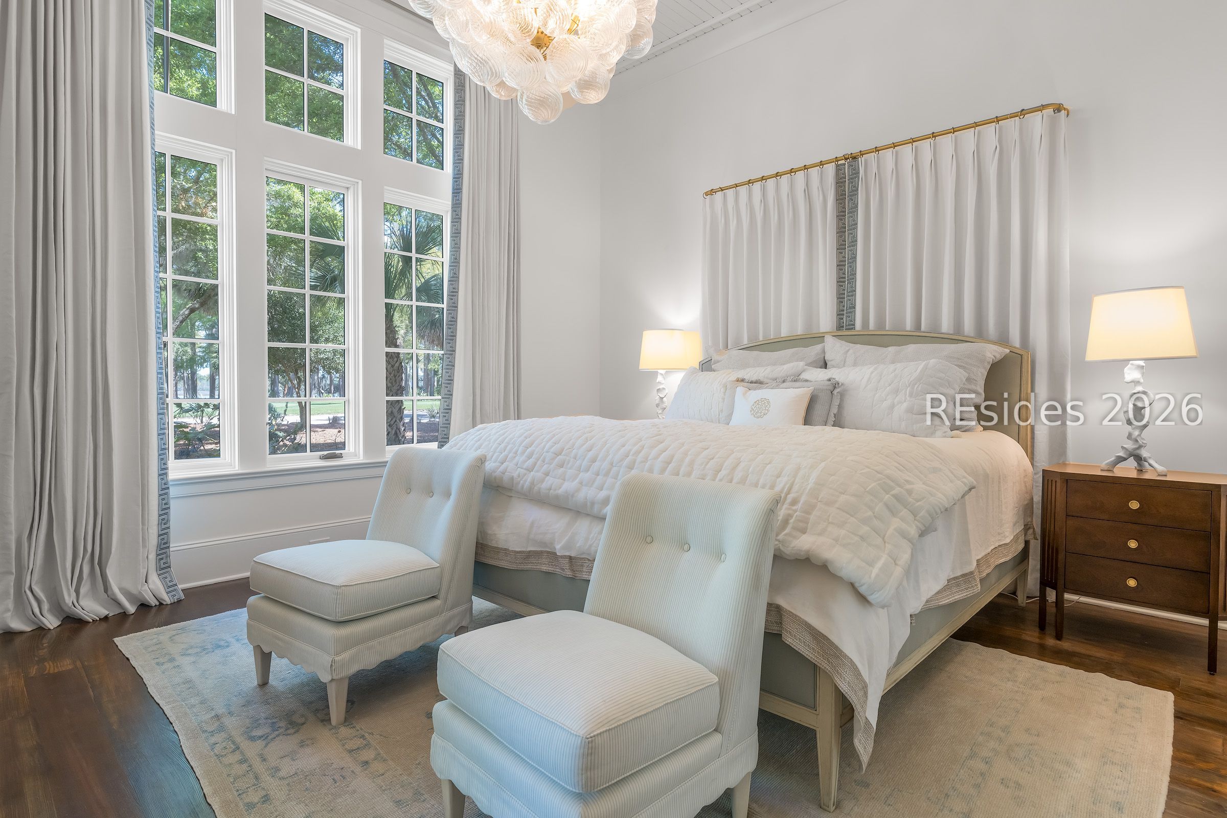 Furnished bedroom with a quilted bed, tall windows, chandelier, and two upholstered chairs.