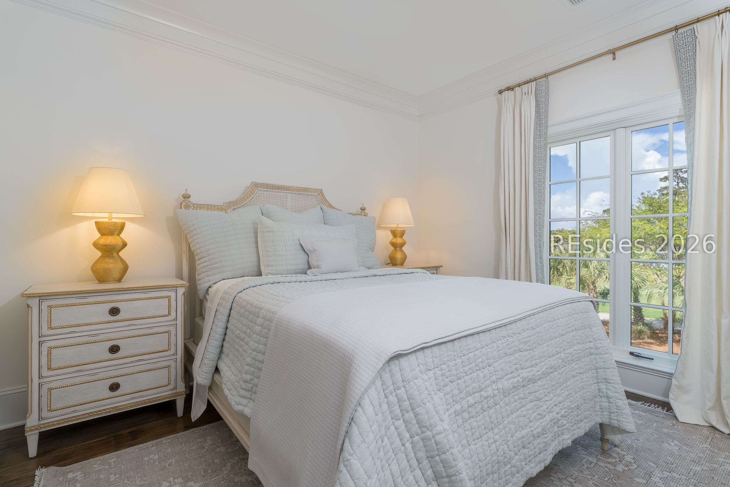Furnished bedroom with a white quilted bed, ornate headboard, and large window overlooking greenery.