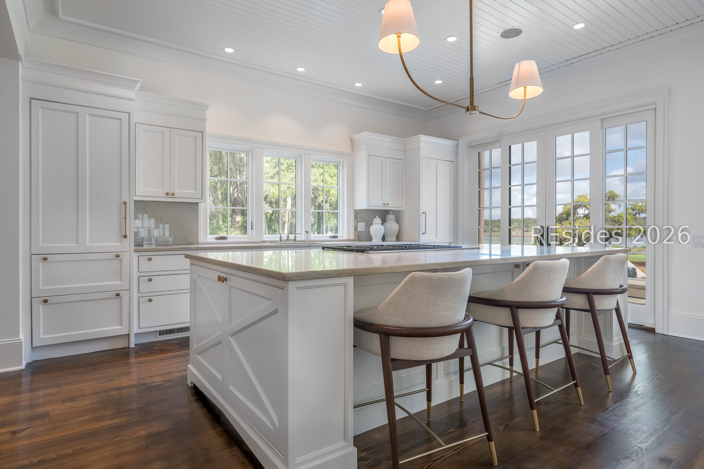 Bright white kitchen with a large island, upholstered stools, and French doors overlooking water.
