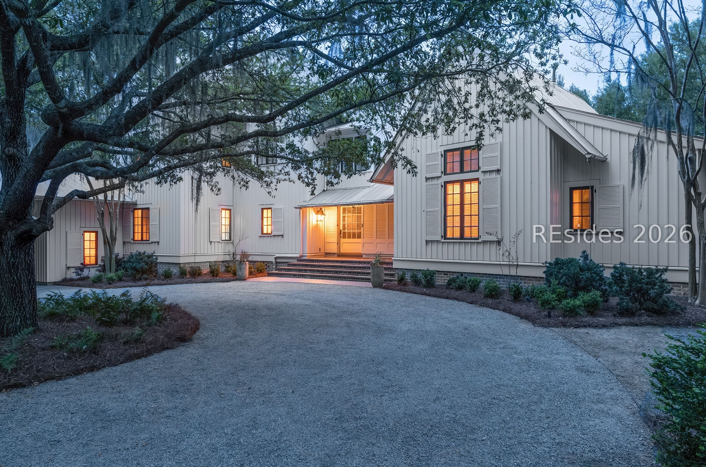 White-sided home with glowing windows, a covered entrance, and a gravel driveway beneath a spreading tree.