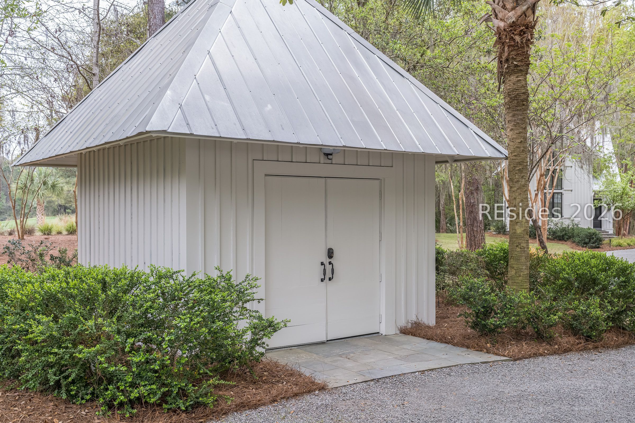 White outbuilding with steep metal roof and double doors among landscaped shrubs.
