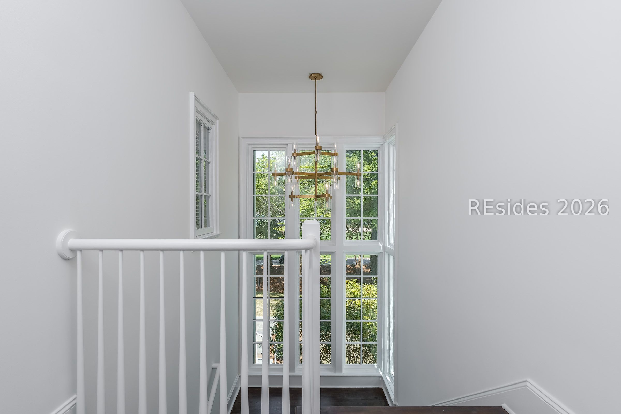 Bright stair landing with white railings, a brass chandelier, and tall windows overlooking trees.