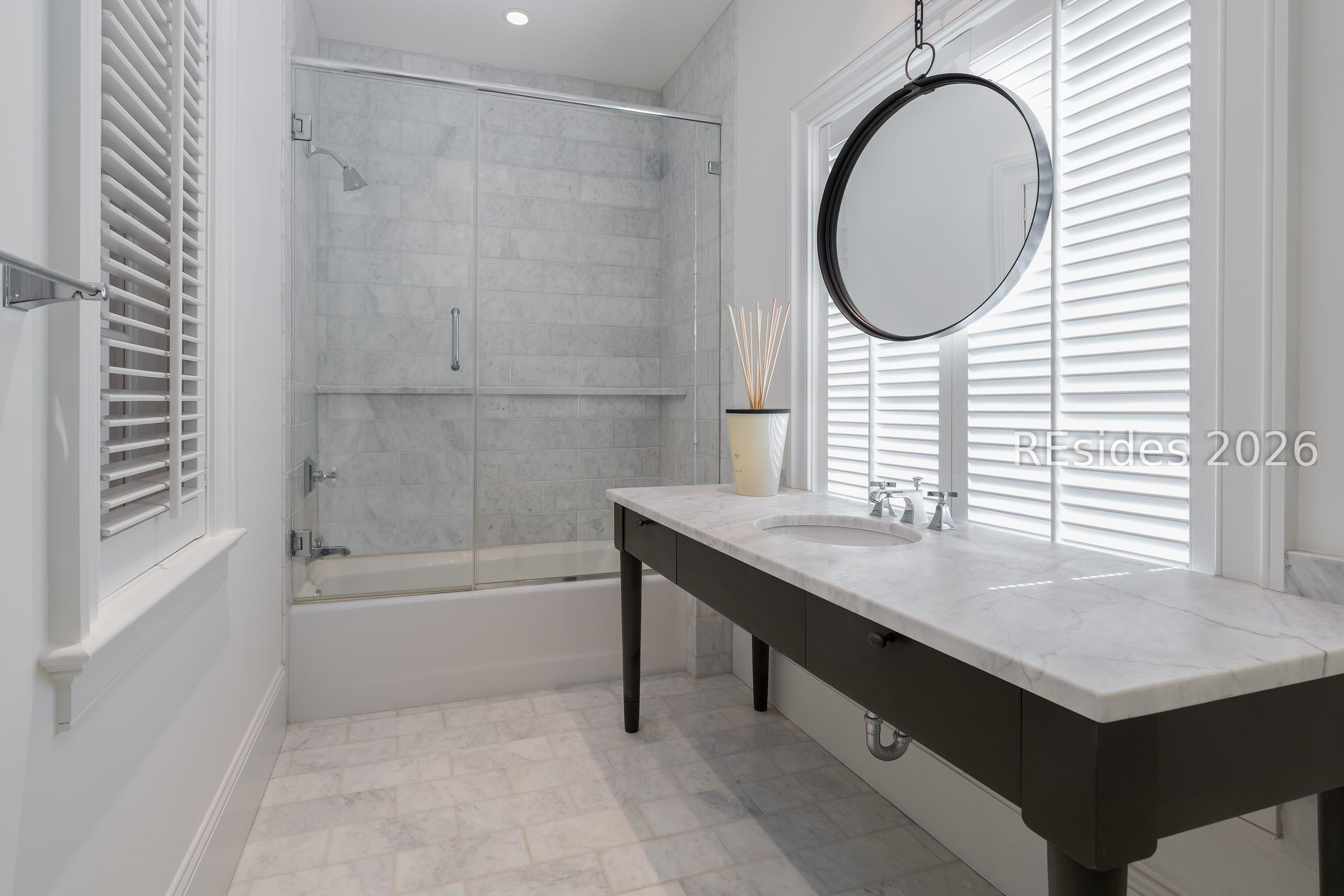 Bathroom with a marble-tile shower, dark vanity, round mirror, and plantation shutters.