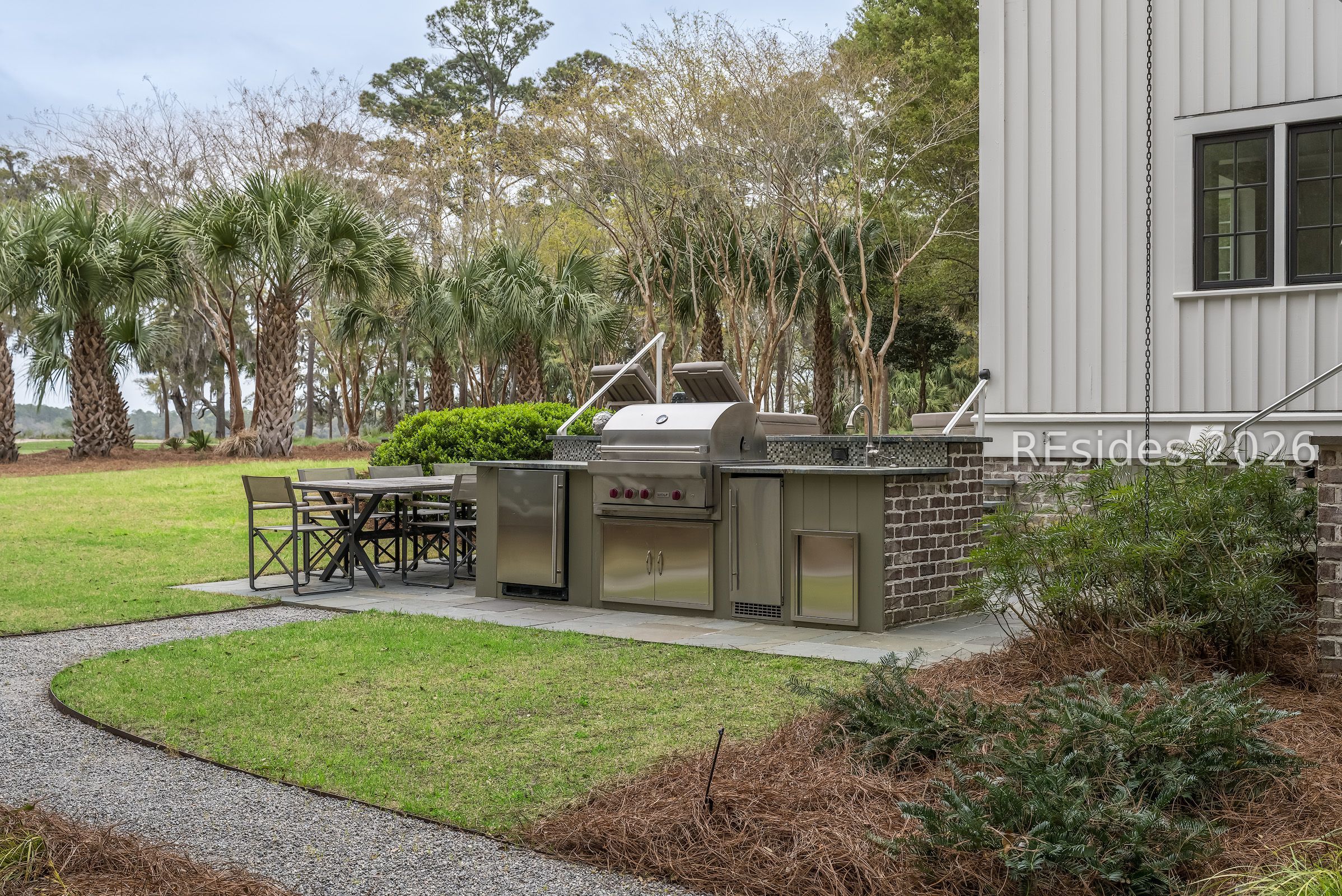Outdoor kitchen with built-in grill and dining table beside the home's landscaped lawn and palms.