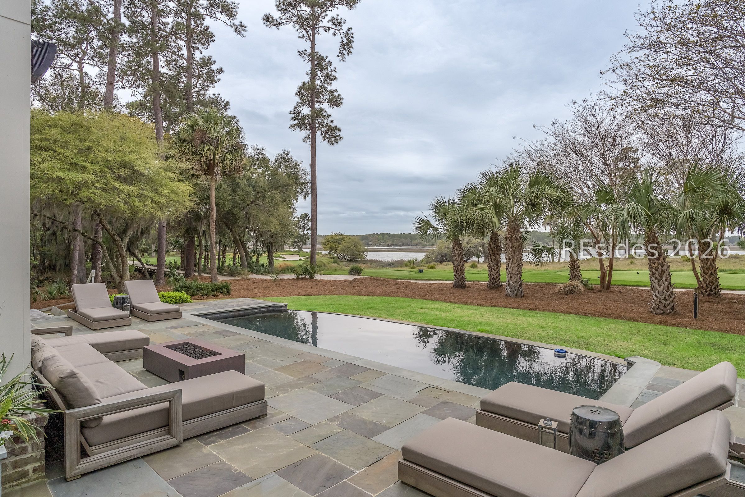 Patio pool with cushioned lounge seating, palms, and views of water and a golf course.