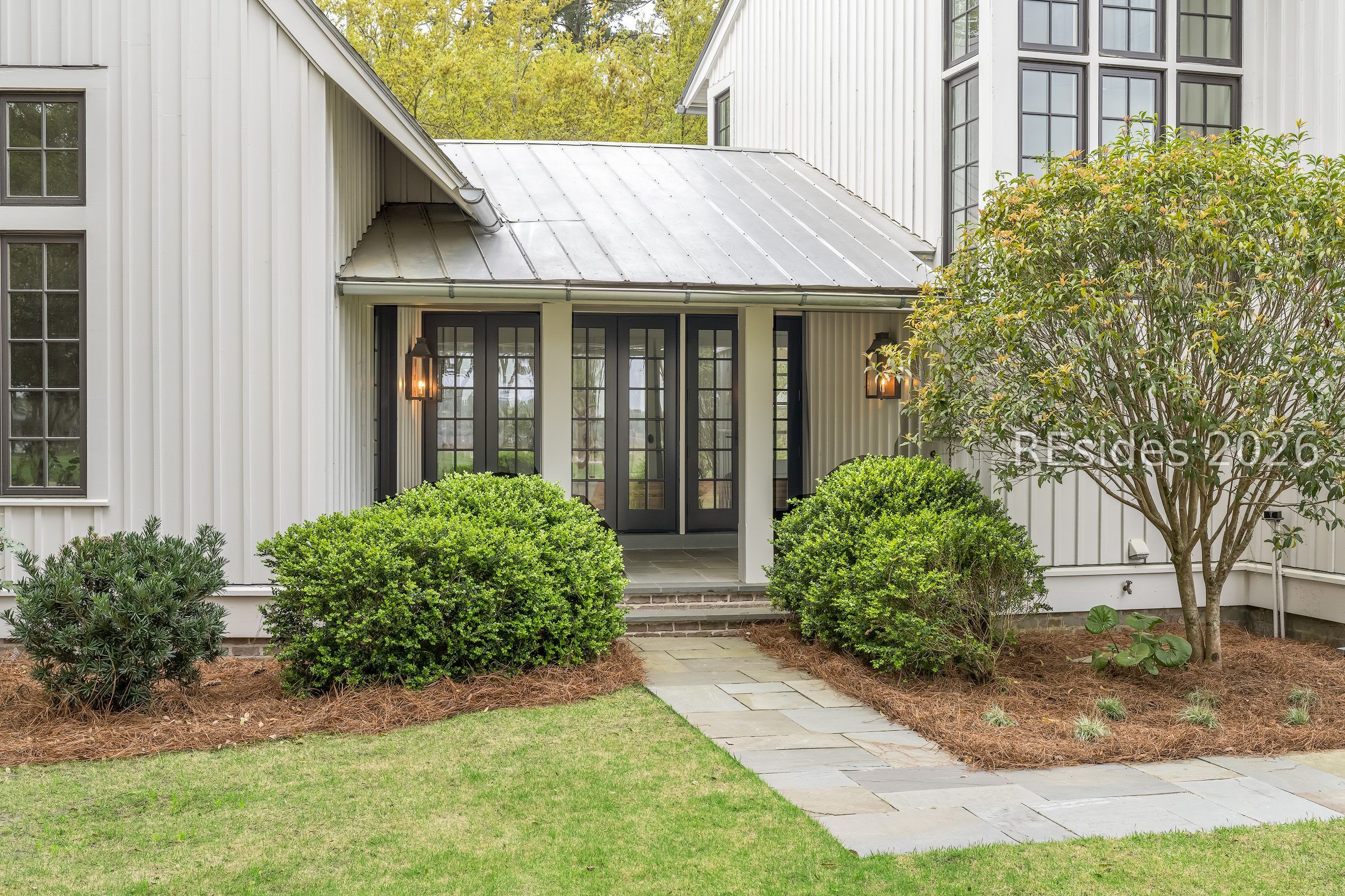 Home entrance with white siding, dark glass-paneled doors, metal roof, and landscaped walkway.