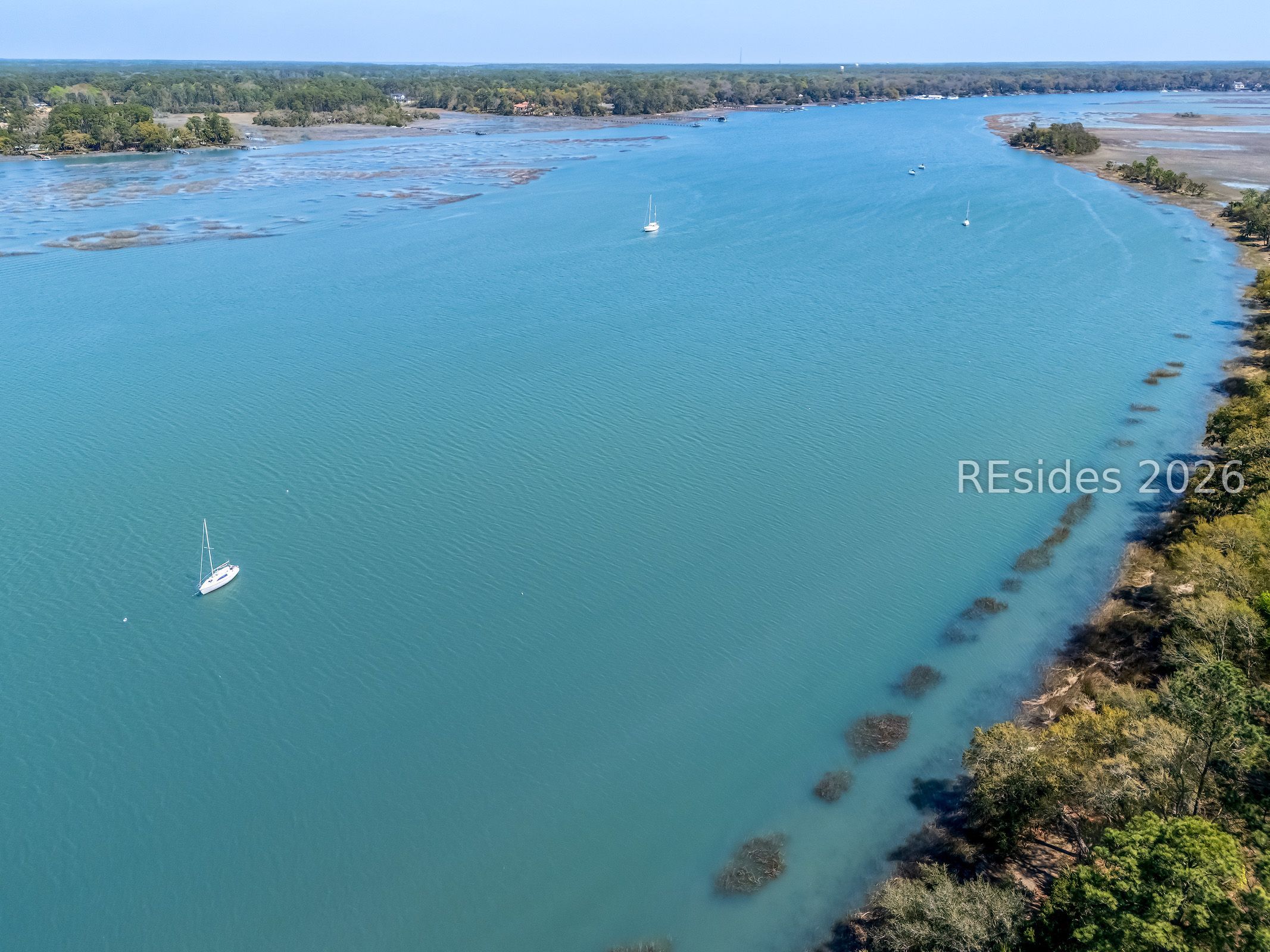 Aerial view across a blue waterway, with sailboats and wooded marsh shoreline.