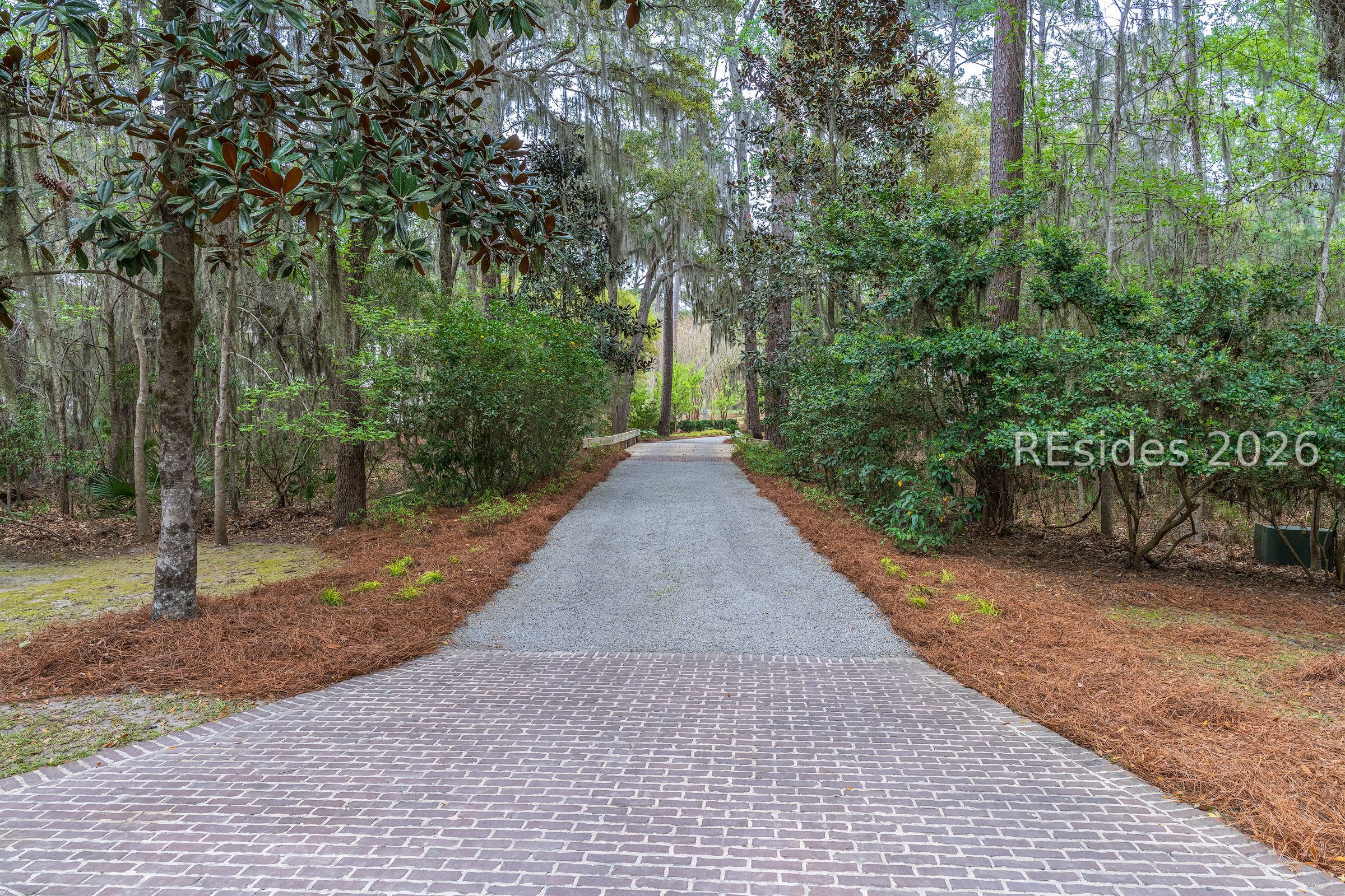 Brick-paved entrance transitions to a gravel driveway beneath mature trees and hanging Spanish moss.