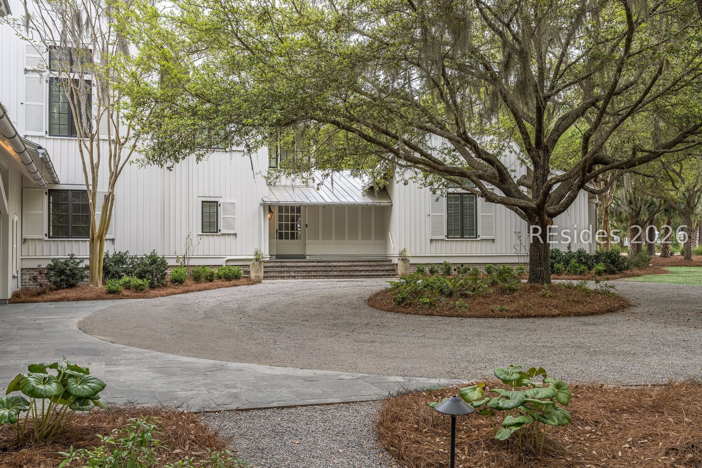 White-sided home with a metal roof and screened porch behind a gravel circular driveway and large oak tree.