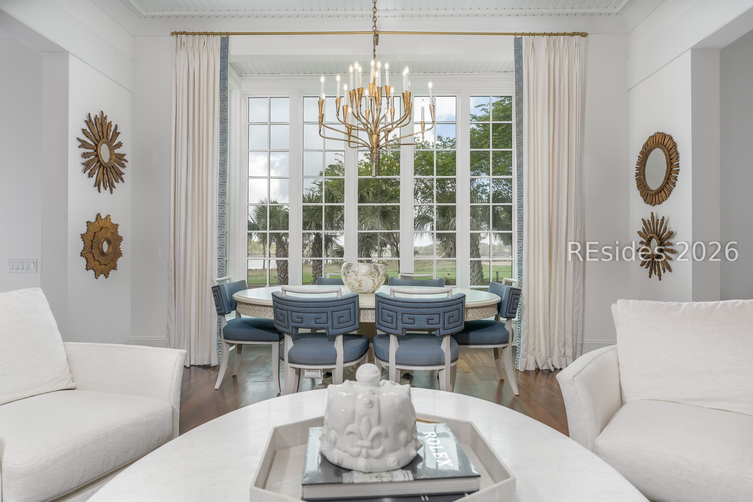 Dining area with a brass chandelier, blue chairs, and tall windows overlooking palms and water.