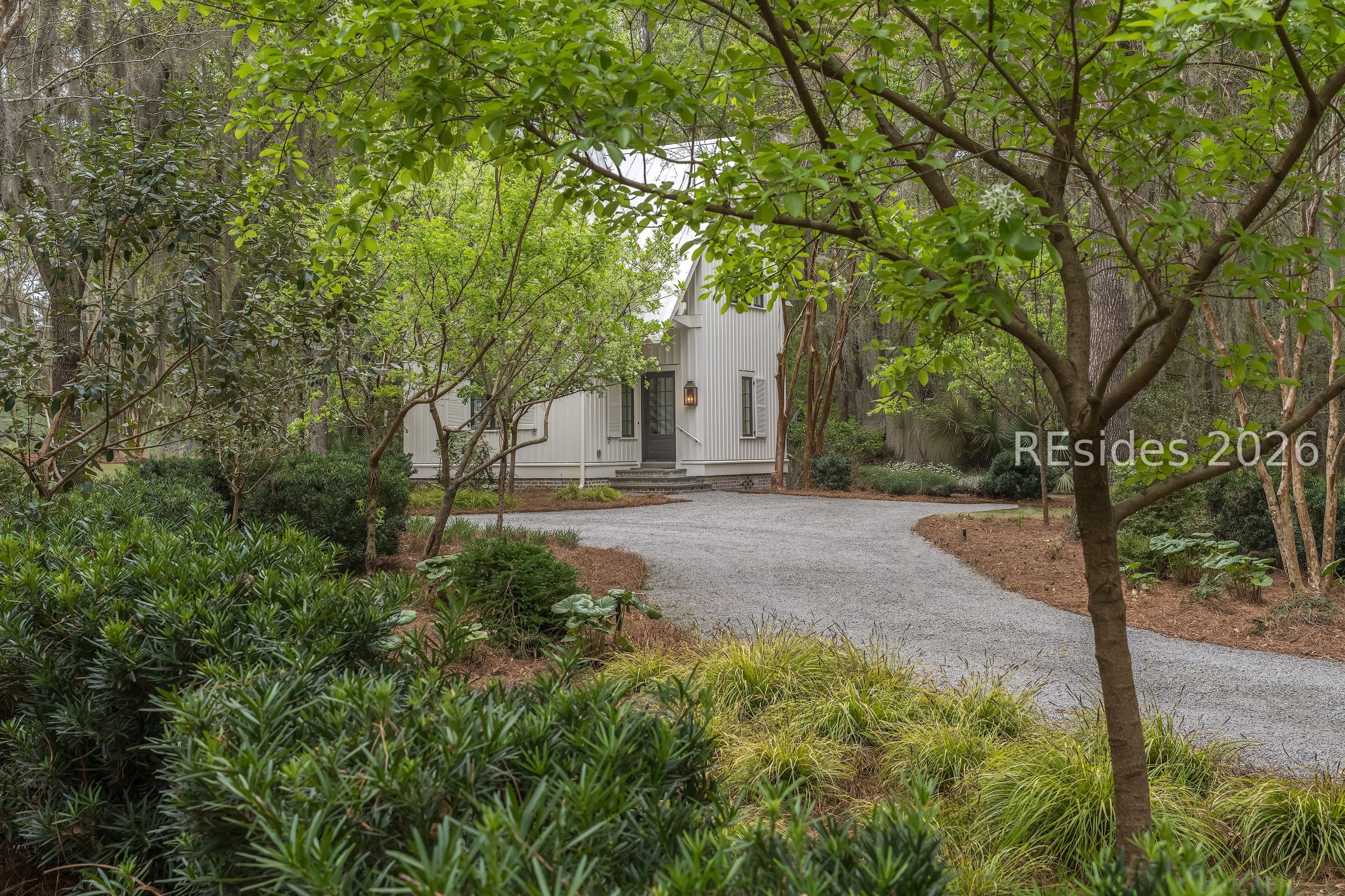 White-sided home framed by mature trees and greenery, with a curved gravel driveway.