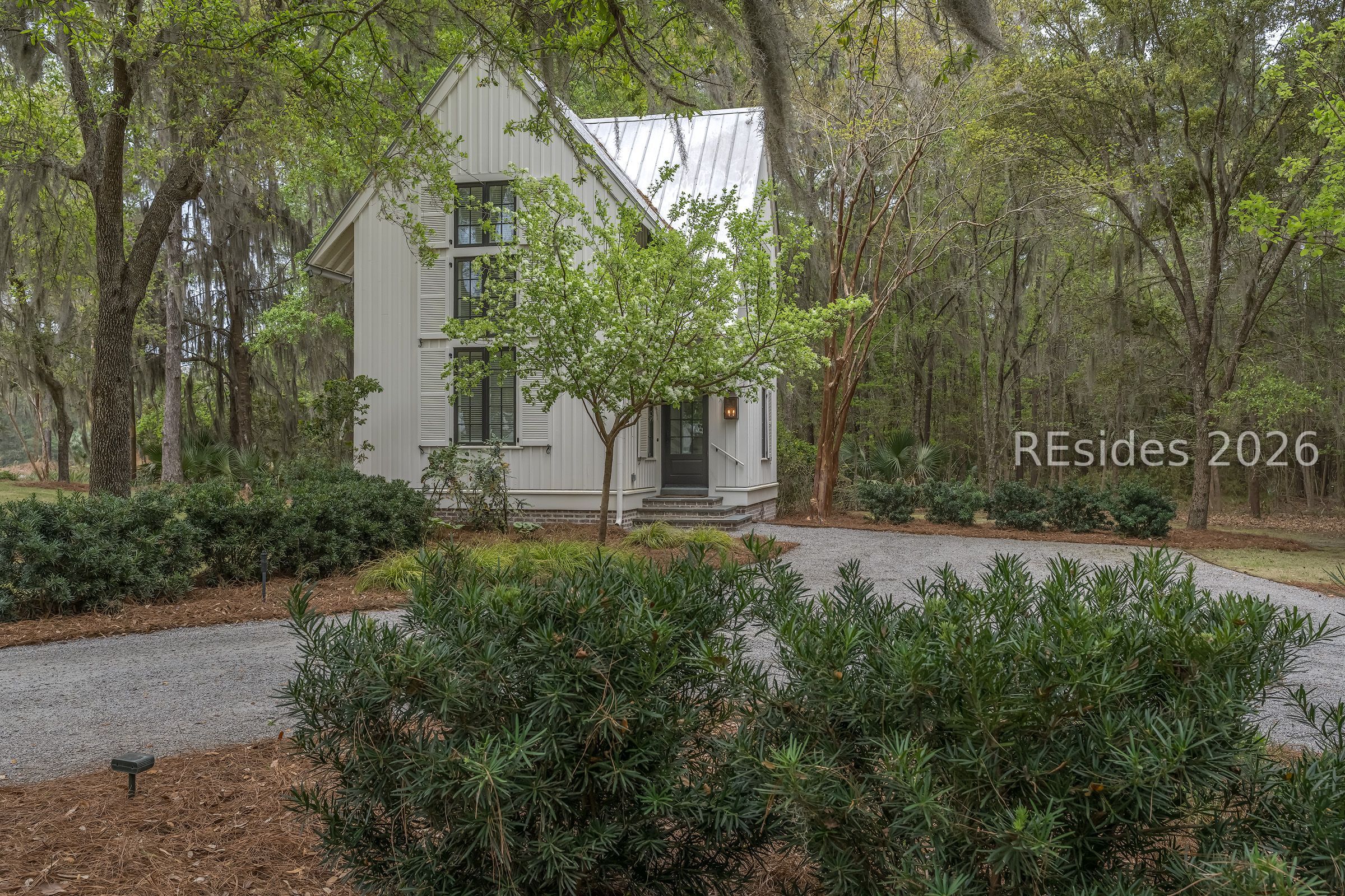 White two-story house with a metal roof, tucked among mature trees beside a gravel driveway.