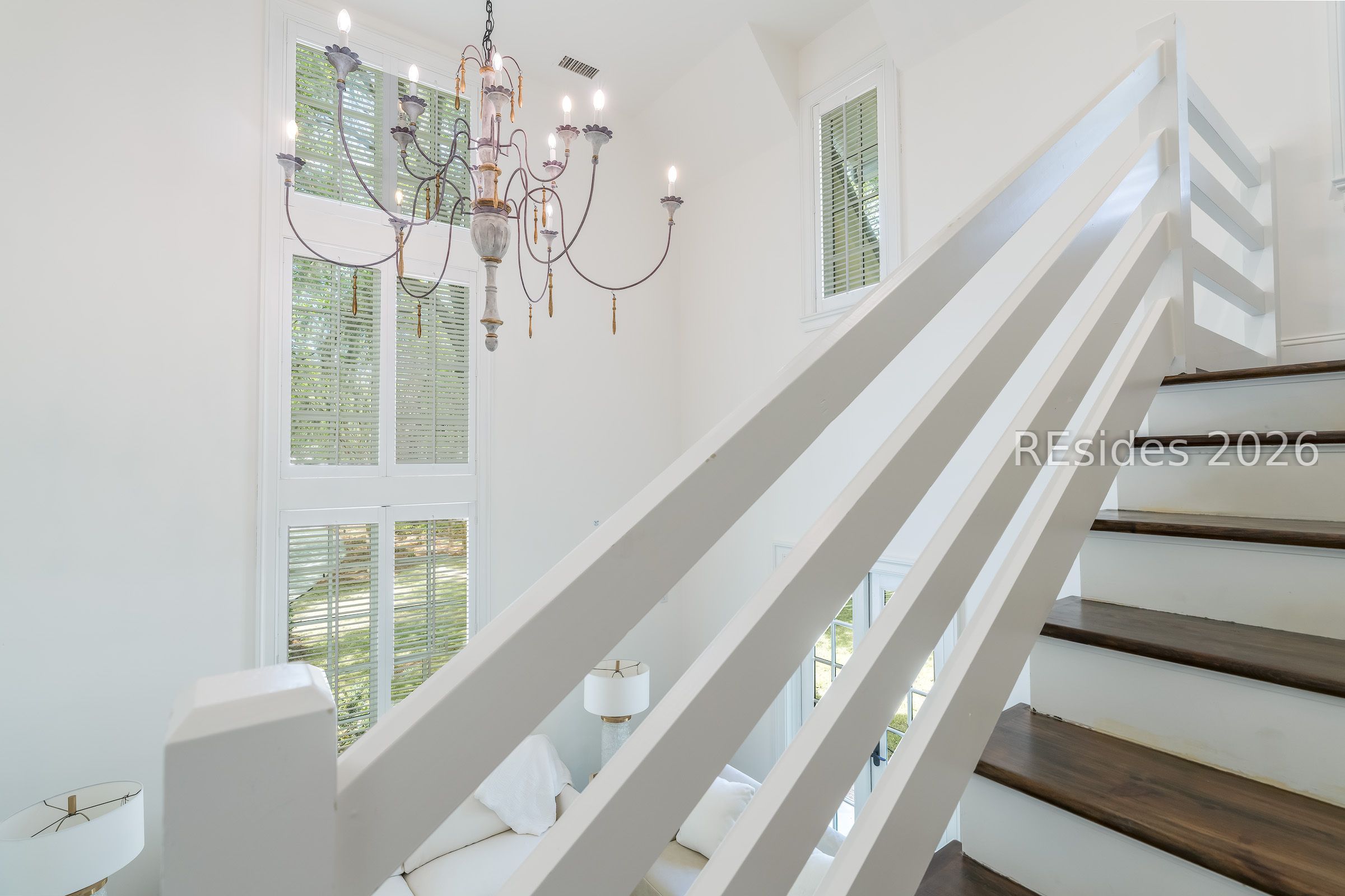Bright stairwell with white railings, dark wood treads, tall shuttered windows, and an ornate chandelier.