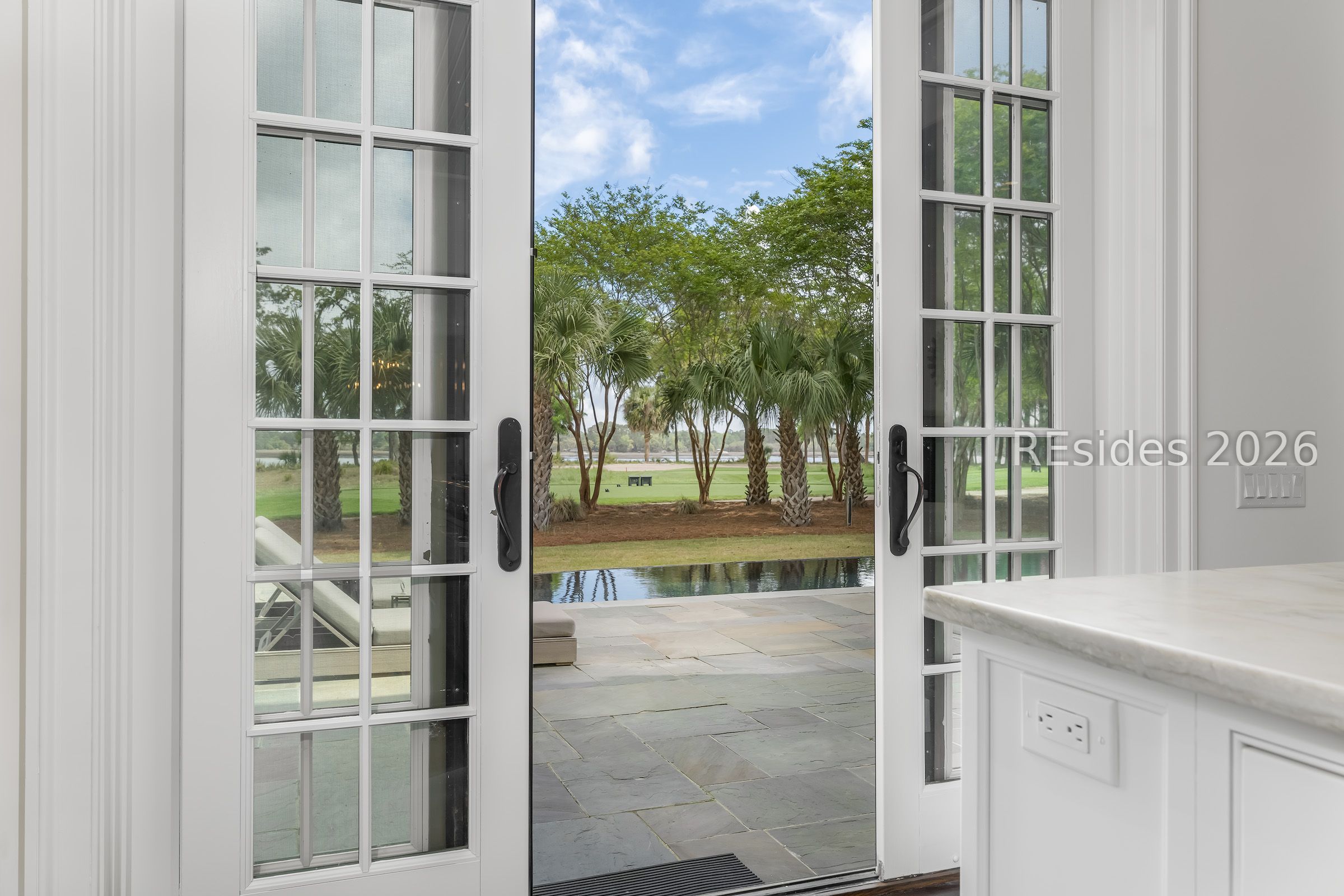 Open French doors frame a stone terrace, reflecting pool, palm trees, and green lawn.