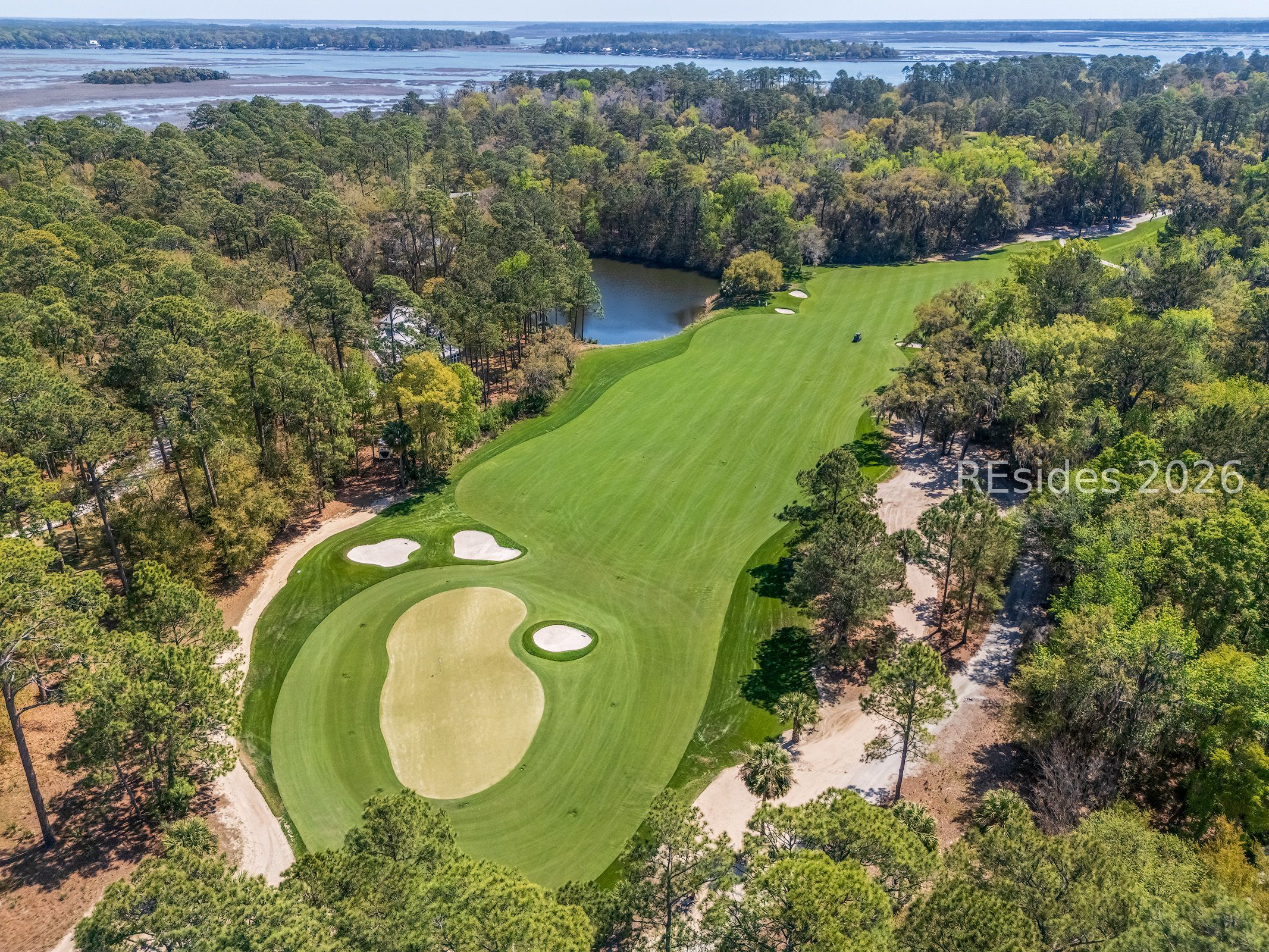 Aerial view of a golf course beside wooded grounds, a pond, and tidal marsh.
