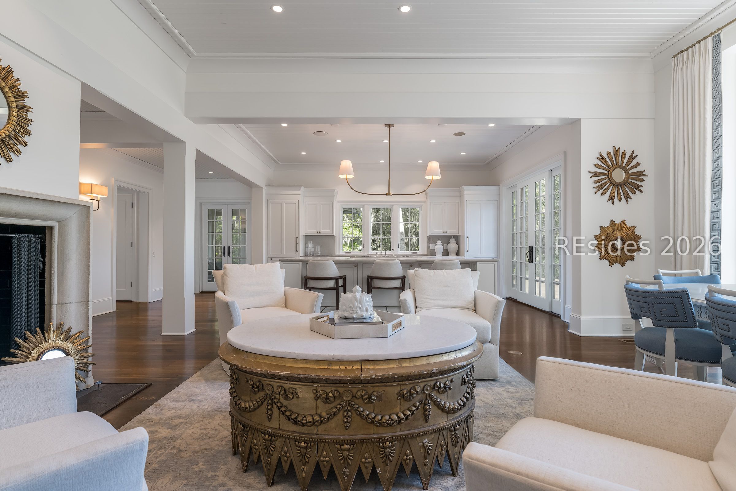 Open-plan living room with white seating, an ornate round coffee table, and a kitchen beyond.