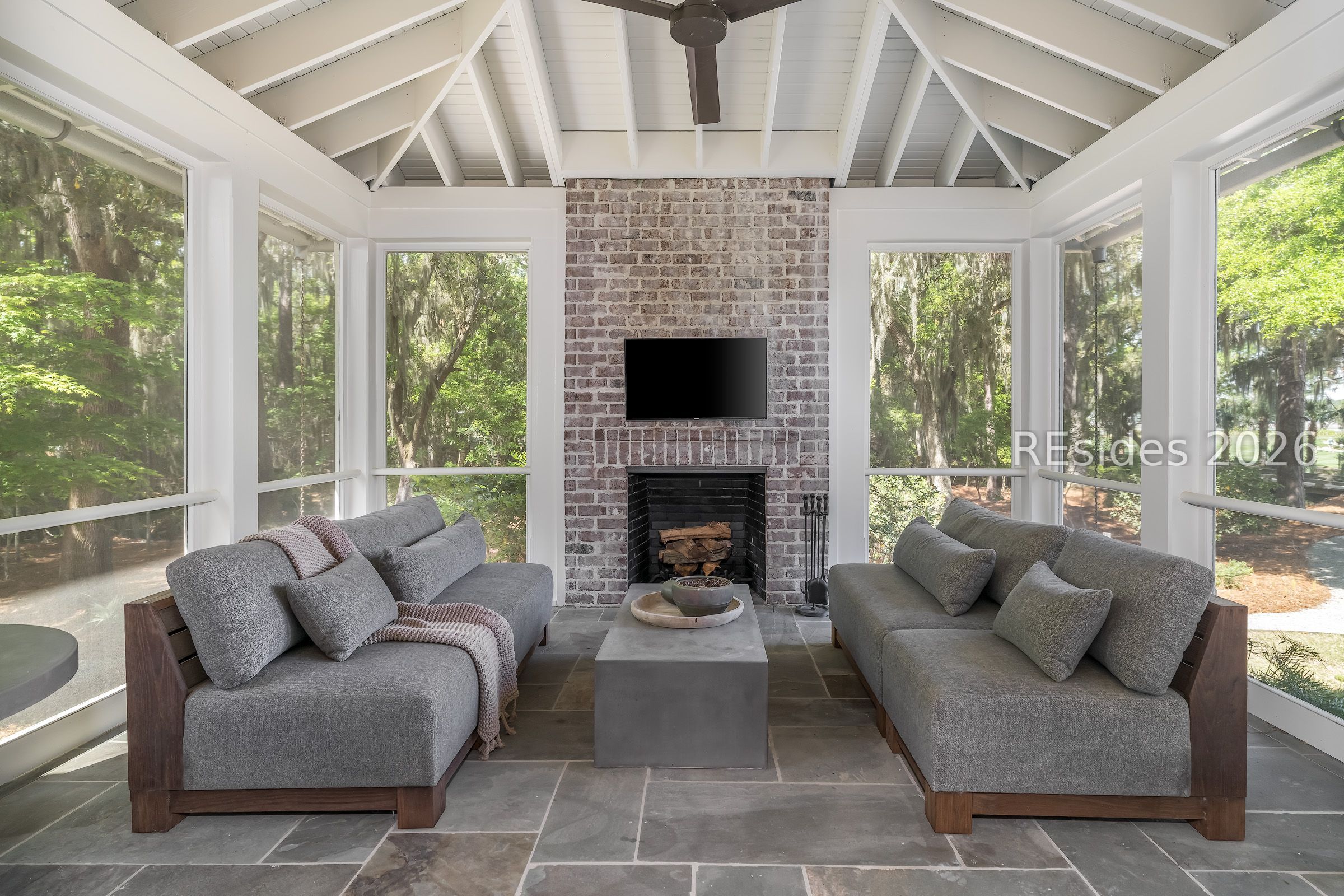Sunroom with gray sofas, a brick fireplace, vaulted ceiling, and large windows overlooking trees.