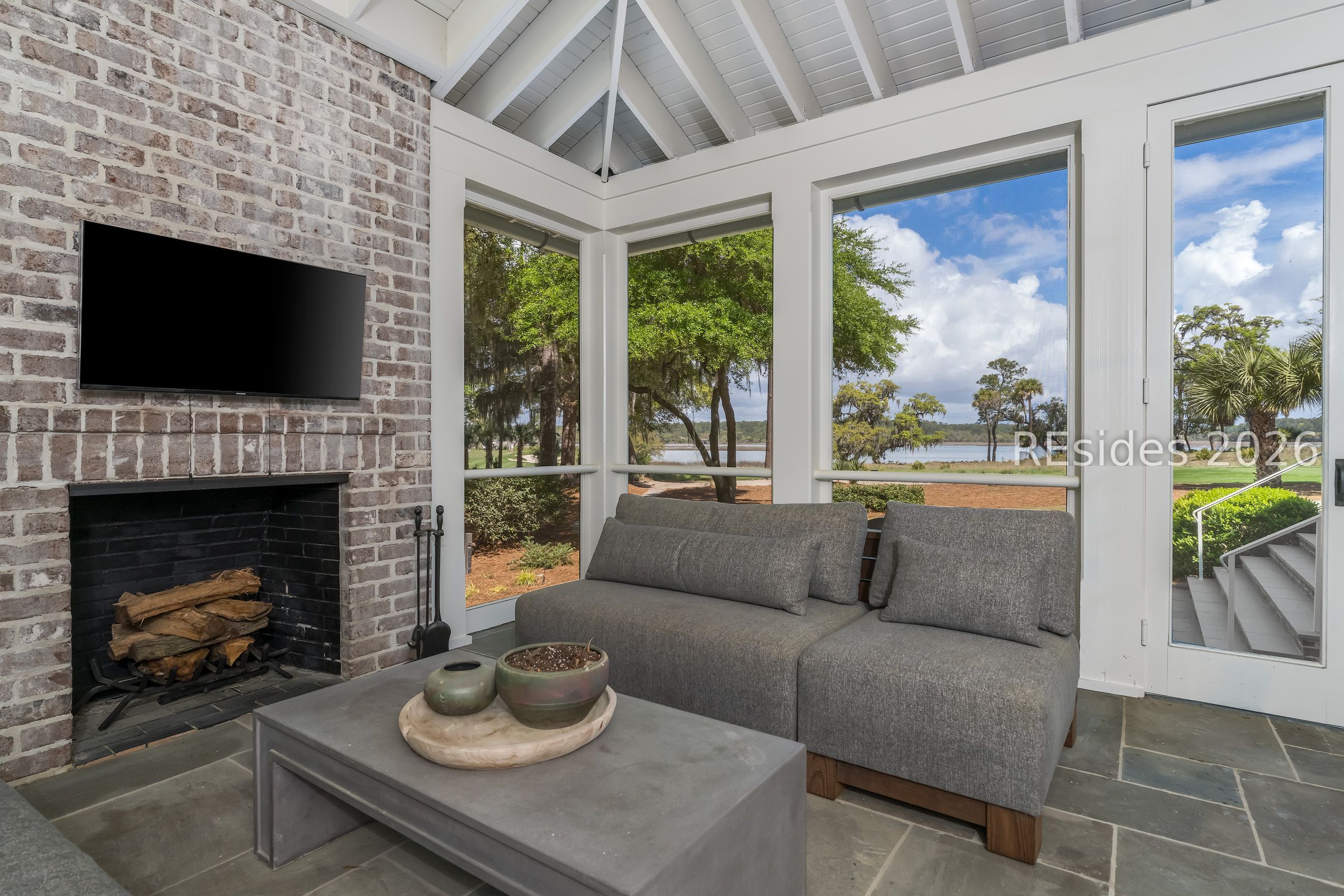 Sunroom with gray sofa and brick fireplace, overlooking trees and water.