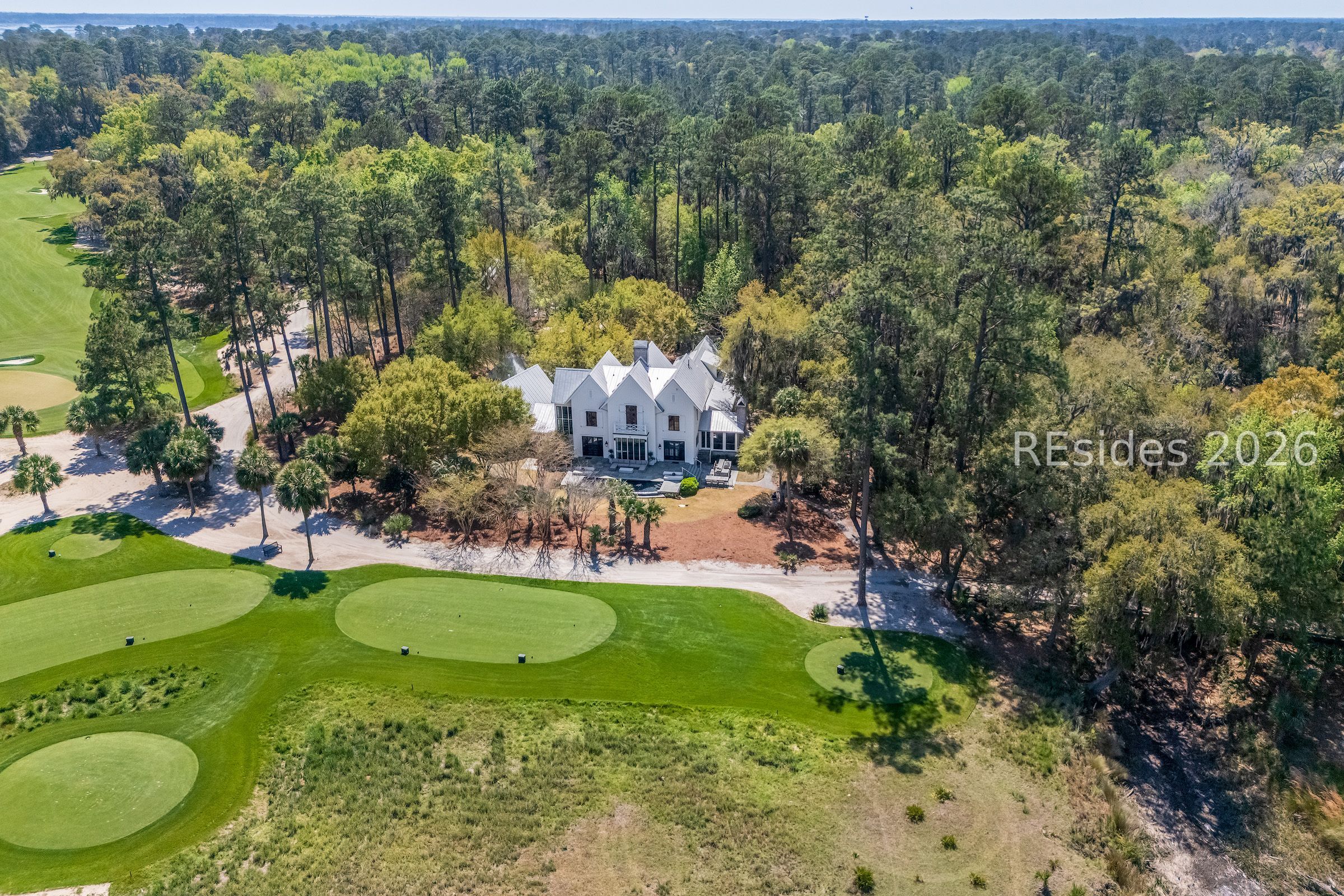 Aerial view of a white gabled home beside golf course greens, surrounded by trees.