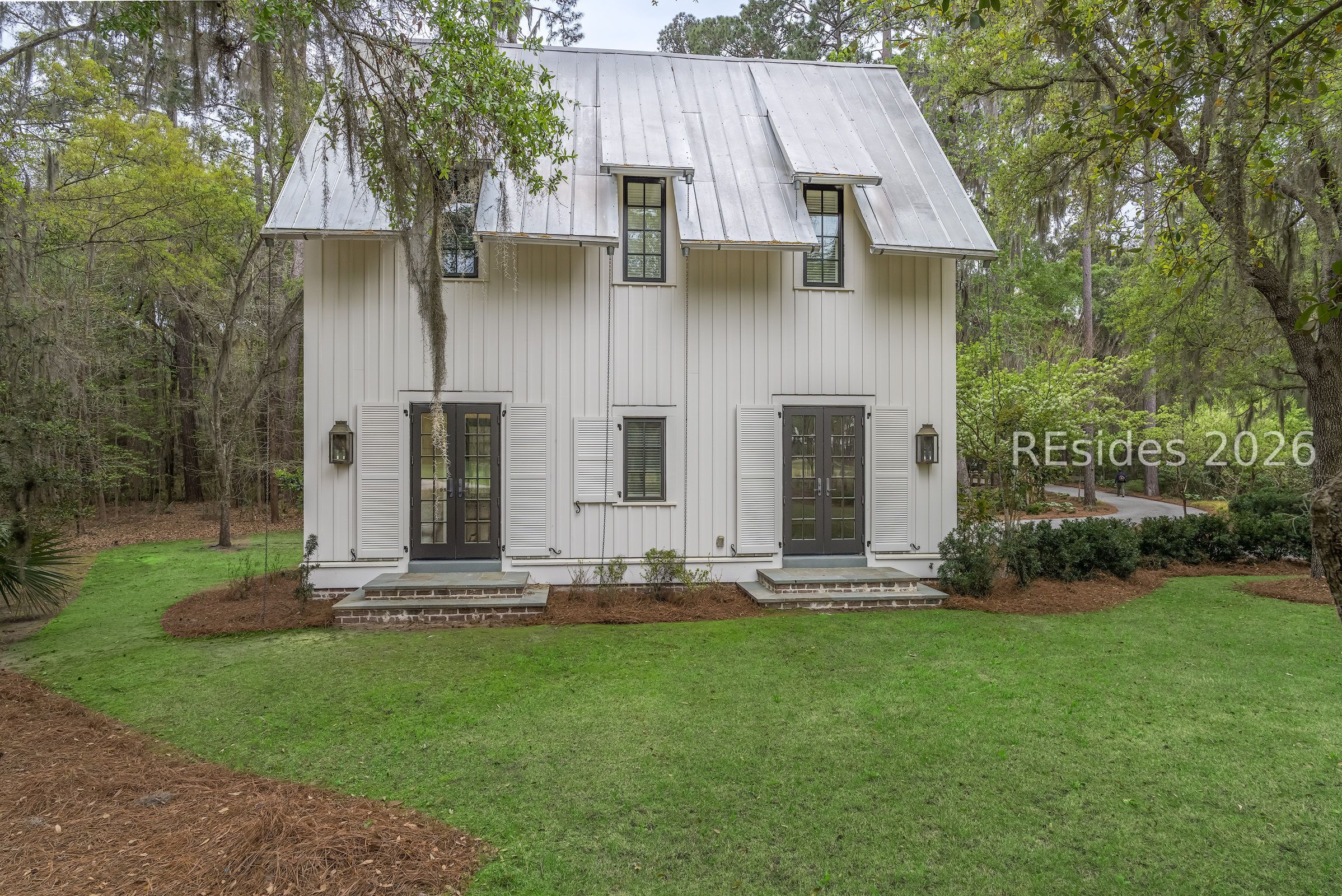 White two-story home with a steep metal roof and twin front entrances, set among mature trees.