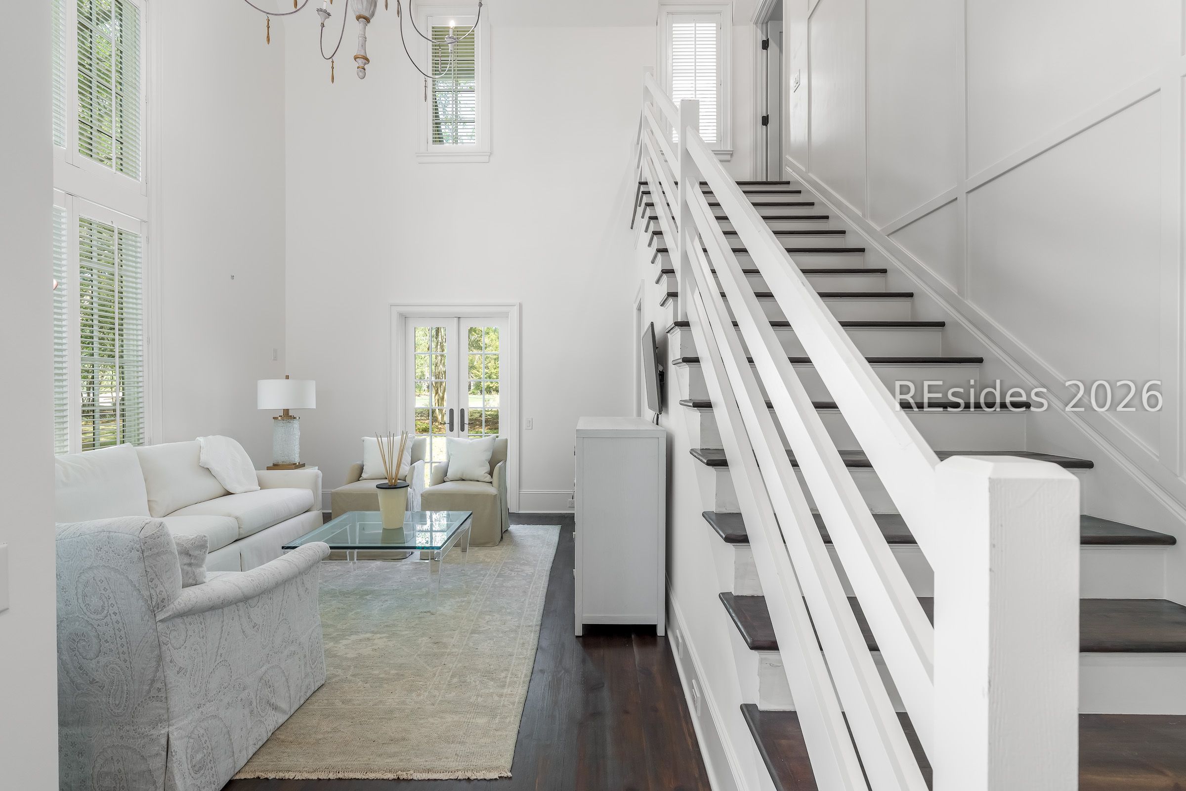 Staged living room with white sofas and a glass coffee table beside a dark-wood staircase.
