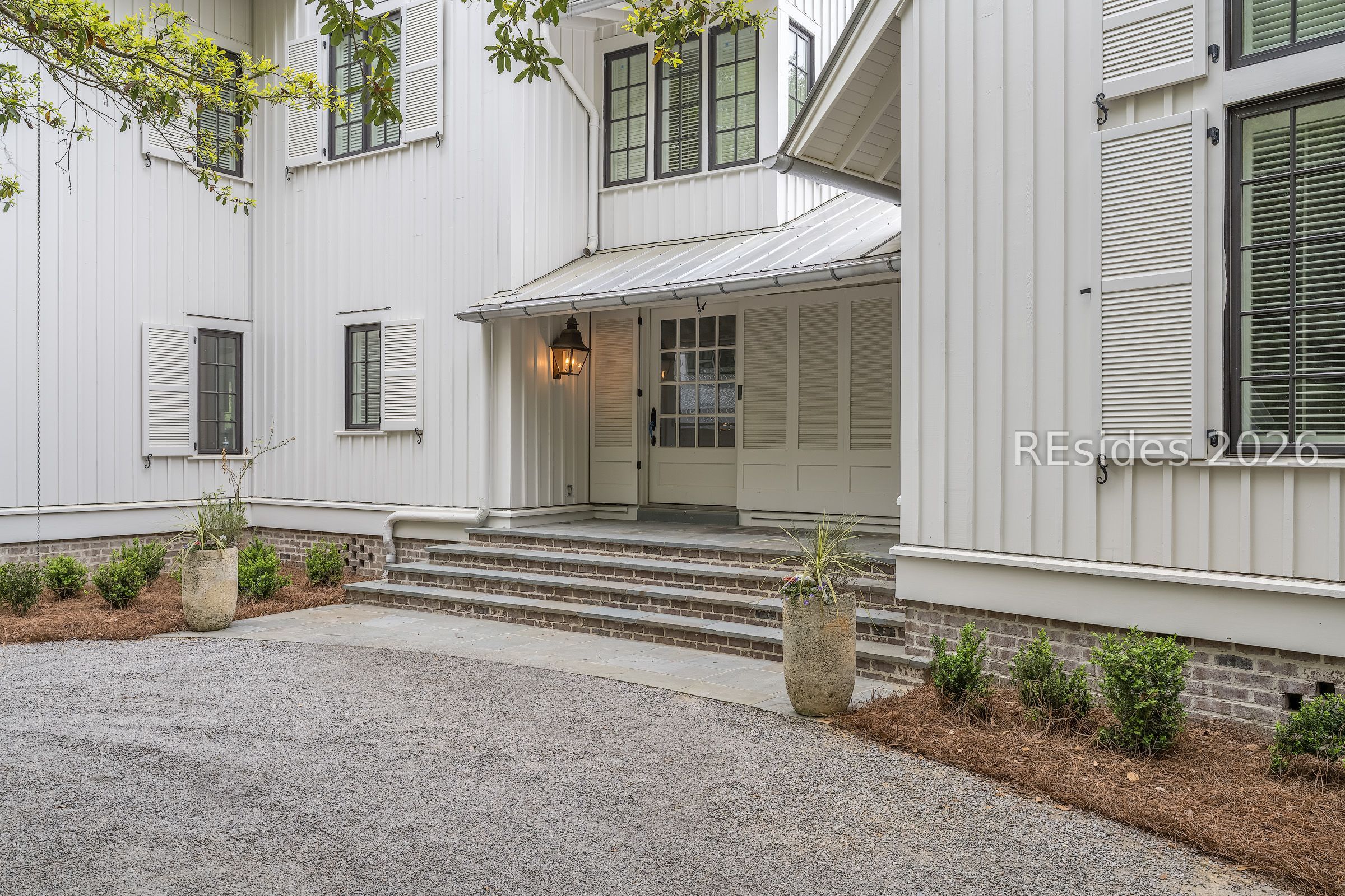 White two-story home entrance with shuttered windows, covered porch, brick steps and gravel drive.