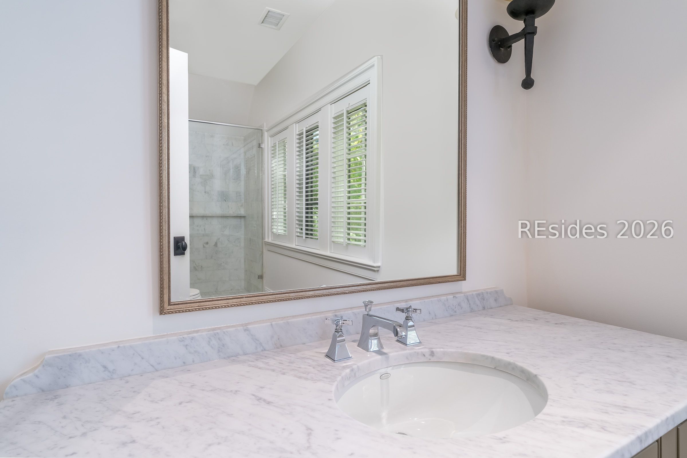 Bathroom vanity with marble countertop and framed mirror reflecting a marble shower and shuttered window.