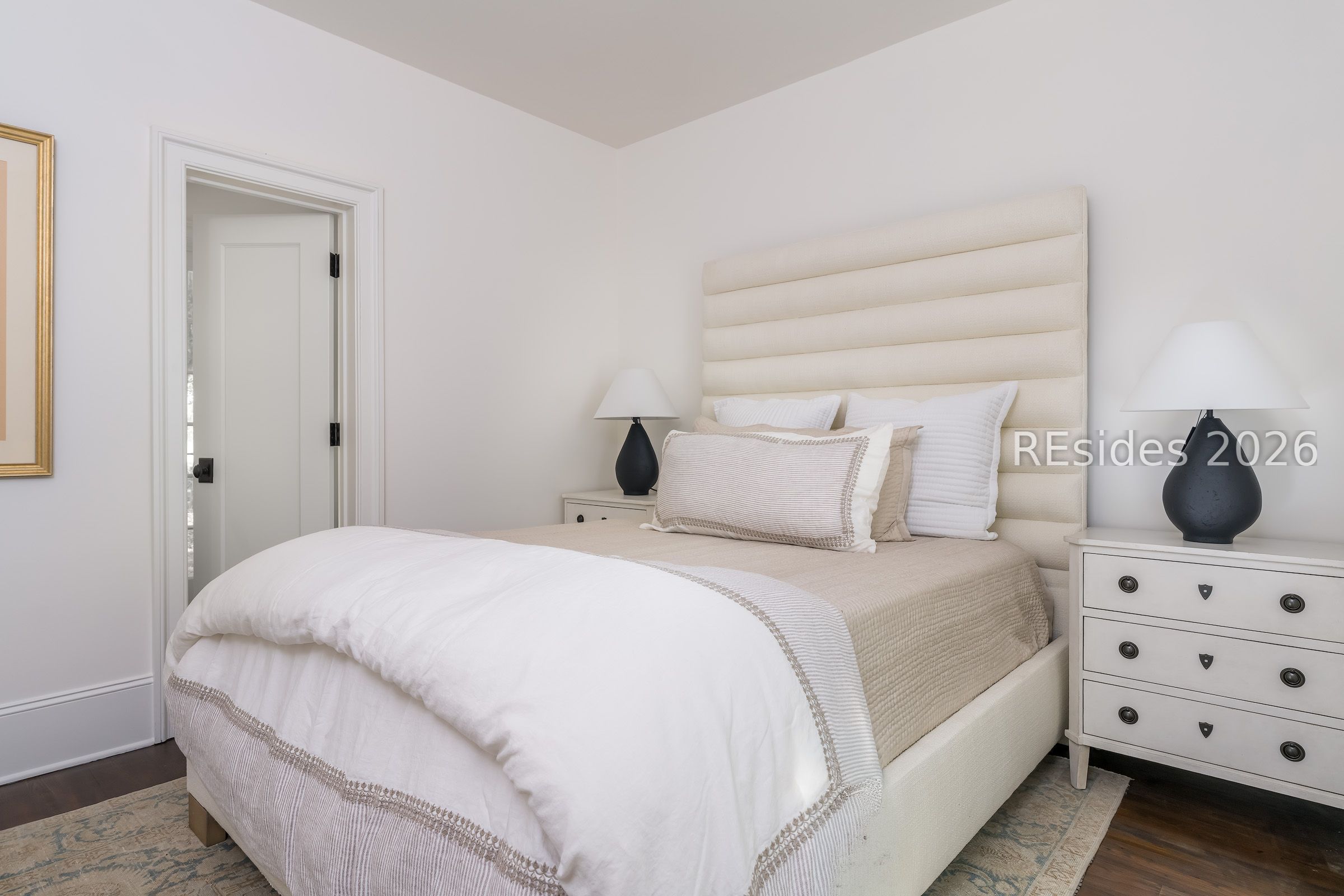 Bedroom with a cream upholstered bed, tall channel-tufted headboard, matching lamps, and a white dresser.