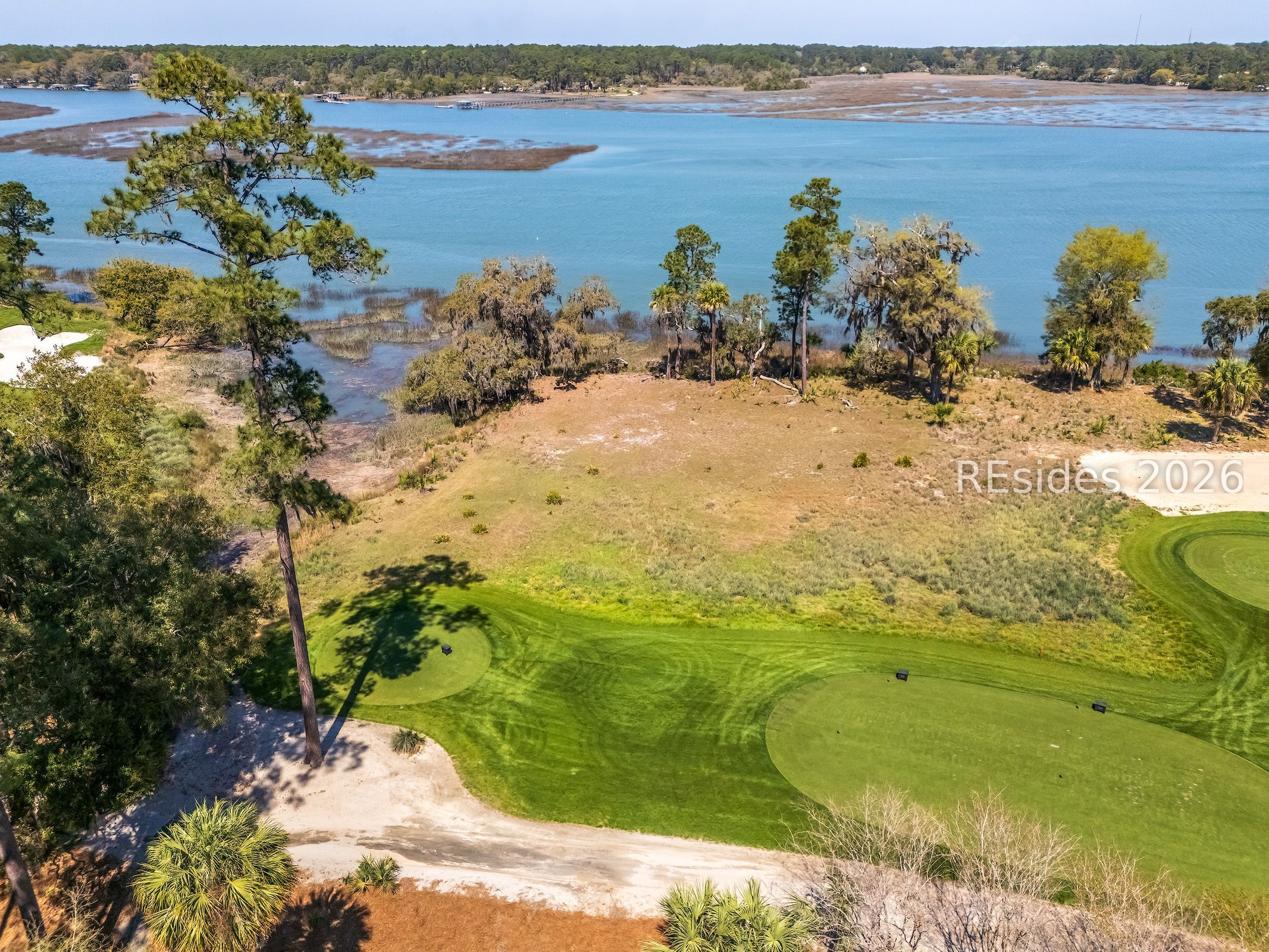 Aerial view of grassy golf greens beside tidal marsh and blue water.