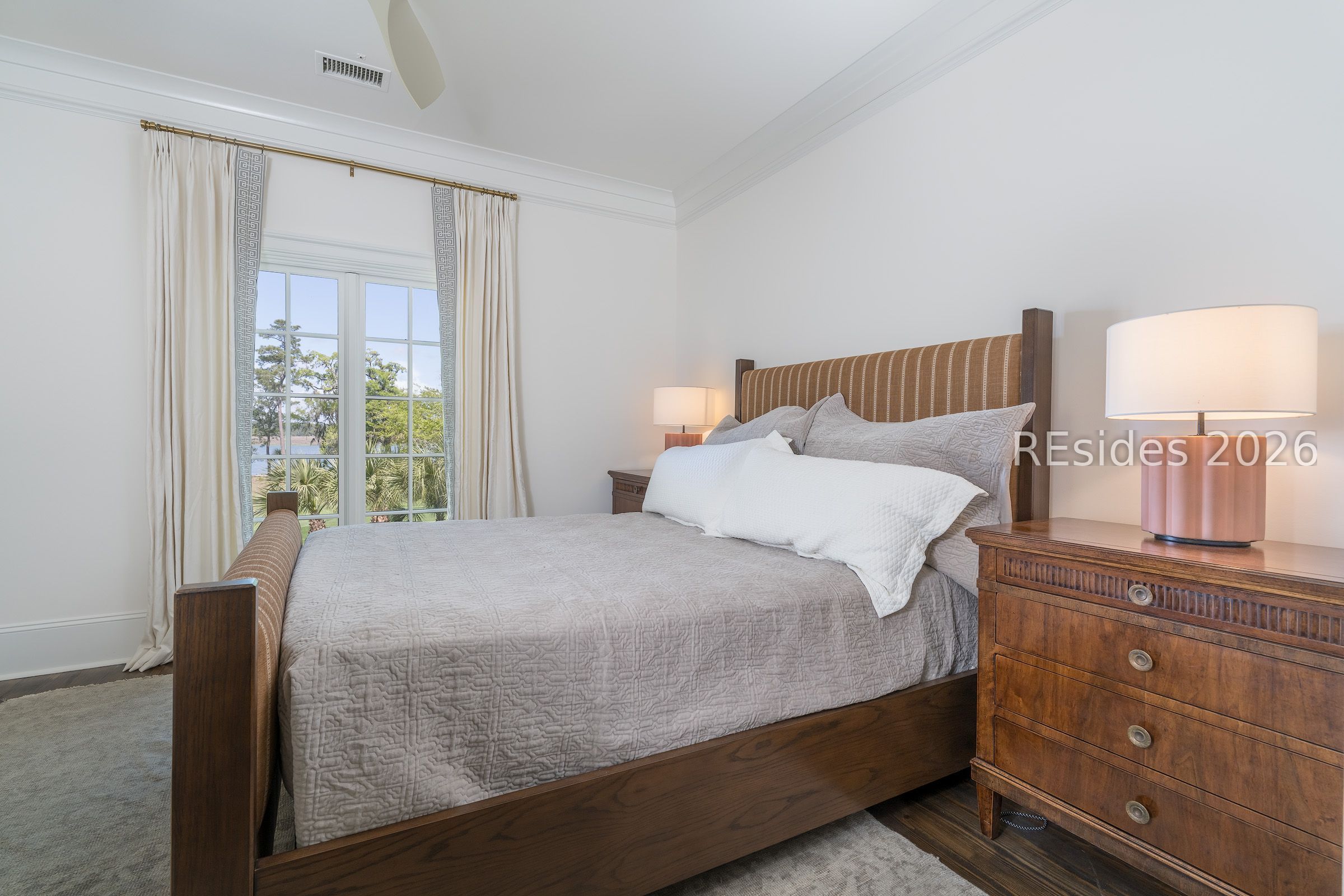 Furnished bedroom with a dark wood bed, gray quilted bedding, and tall windows overlooking trees.