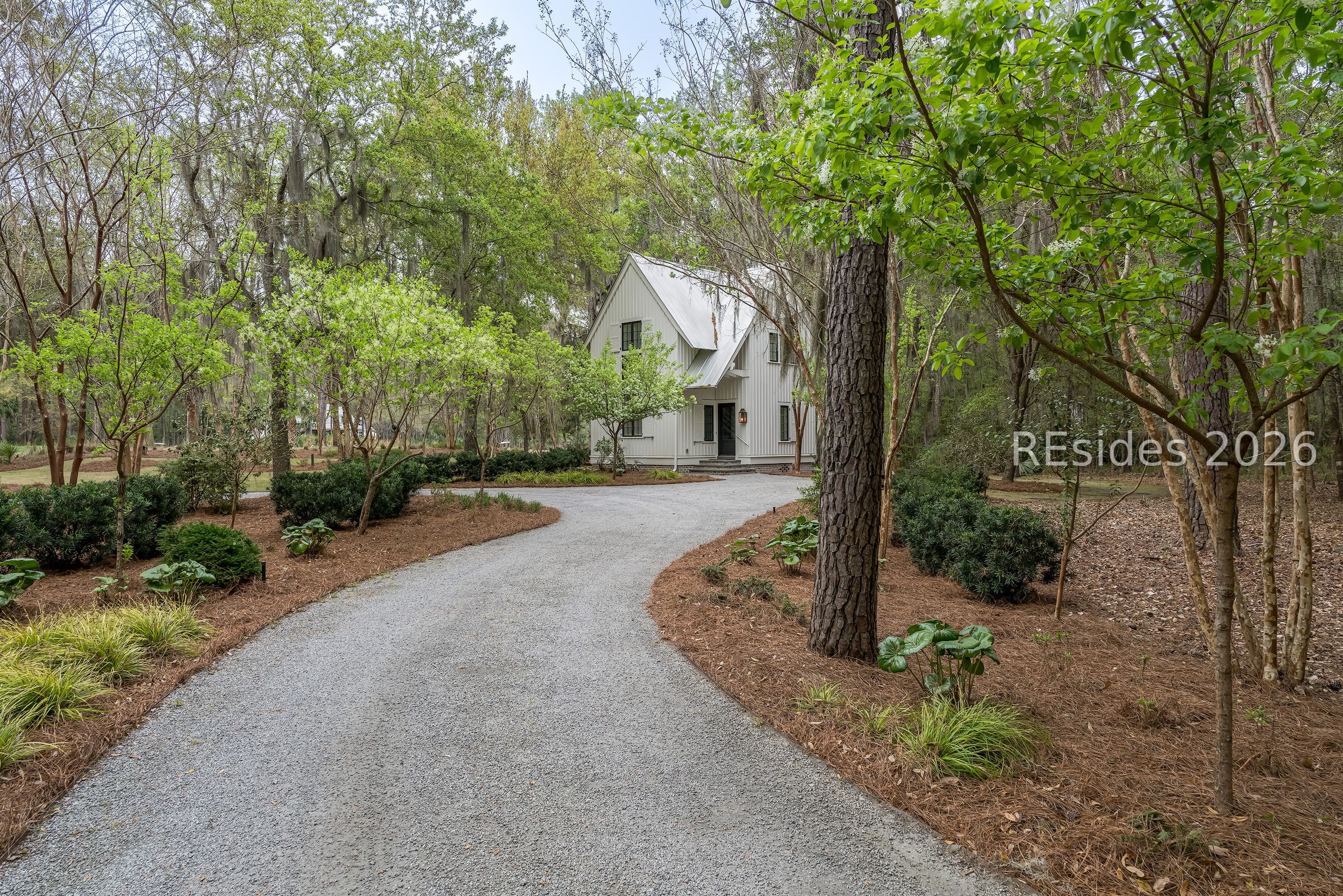 White gable-front house surrounded by trees, with a curving gravel driveway.