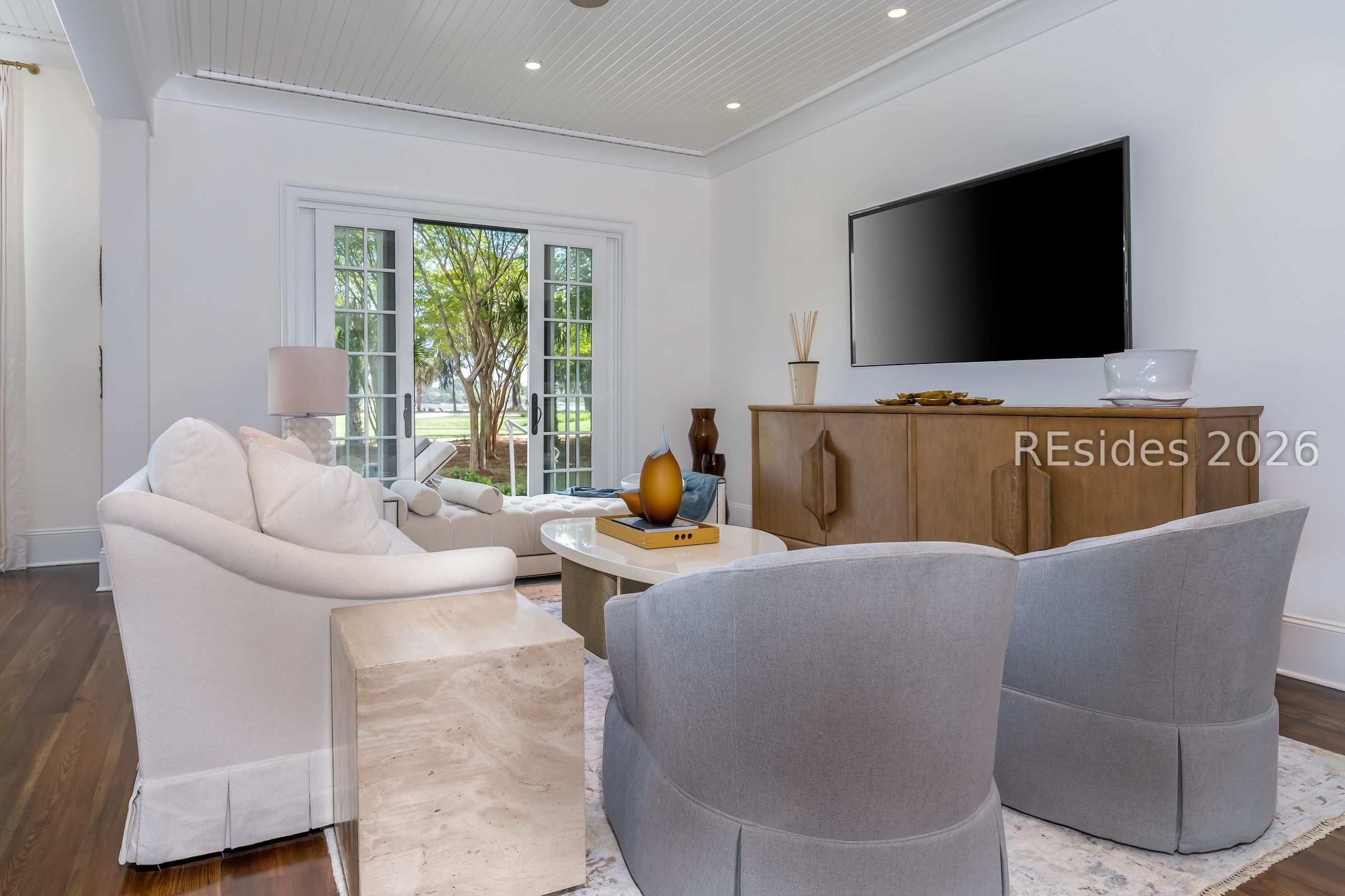 Furnished living room with French doors overlooking a leafy yard and a large television above a wood console.