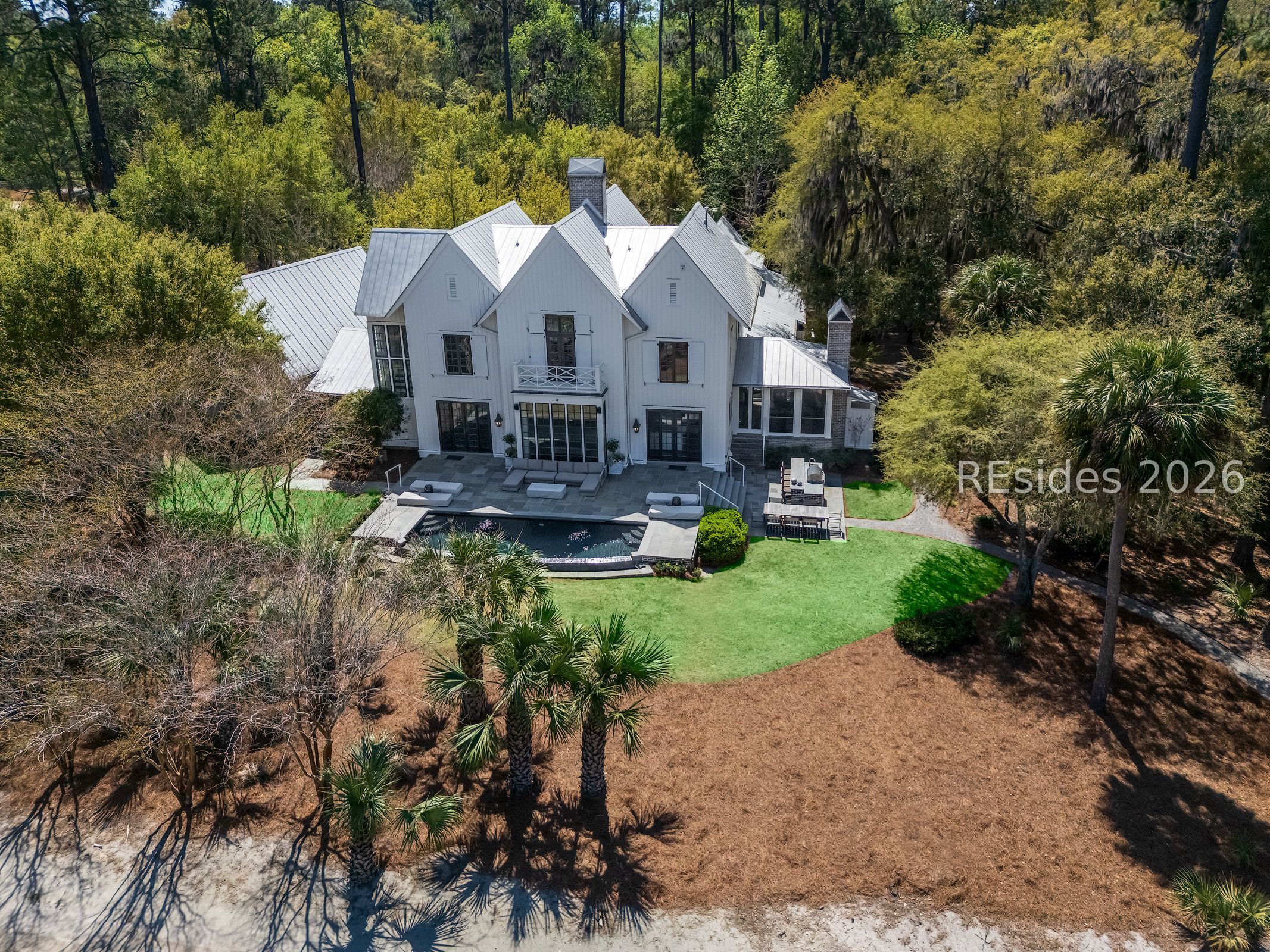 Aerial view of a white home with a metal roof, backyard pool, patio, and wooded grounds.