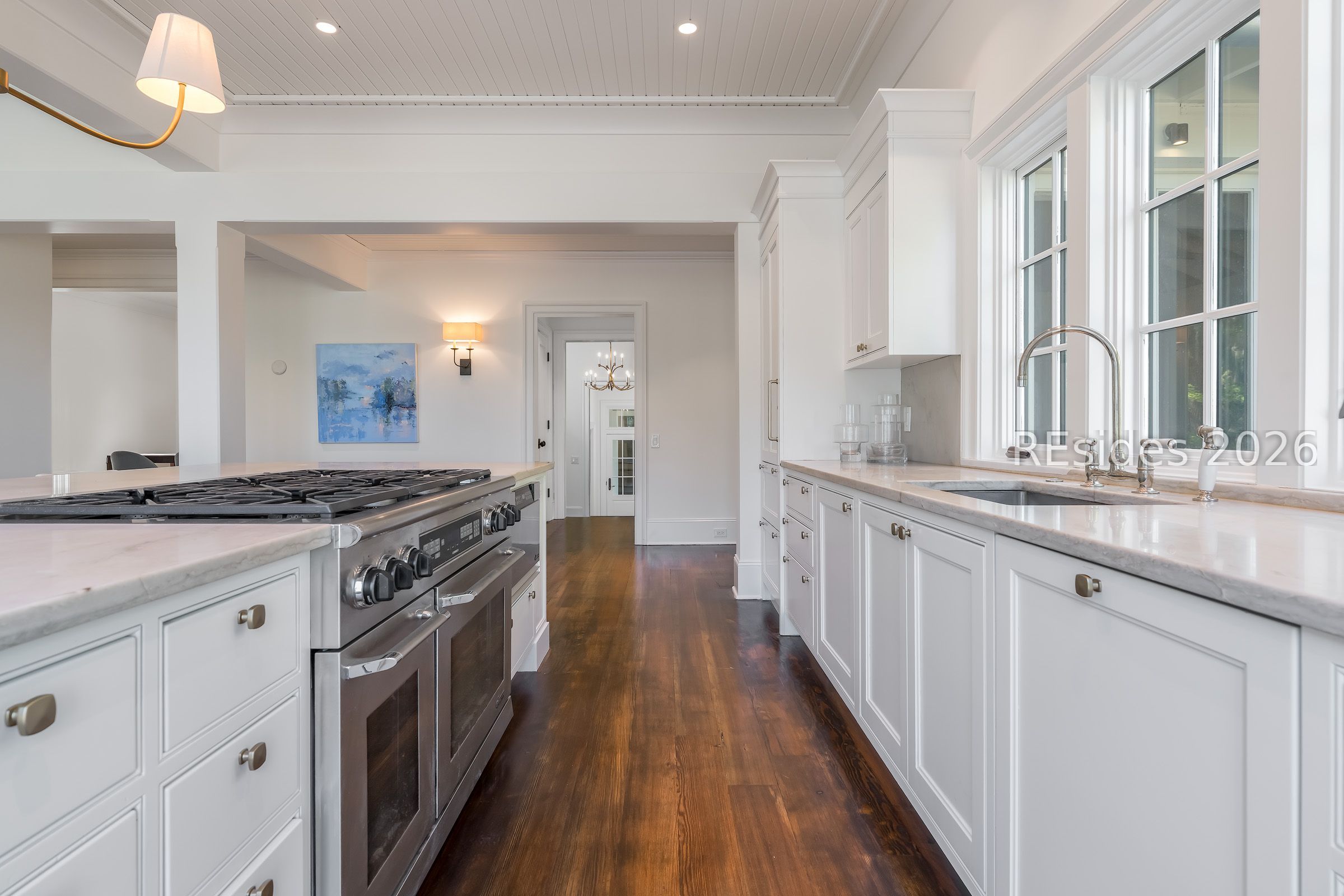 Bright white kitchen with marble counters, a large stainless steel range, and tall windows above the sink.