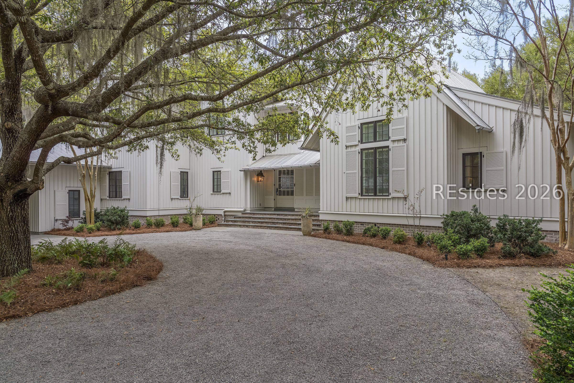 White board-and-batten house with a metal roof, shuttered windows, and a gravel driveway beneath a sprawling oak.