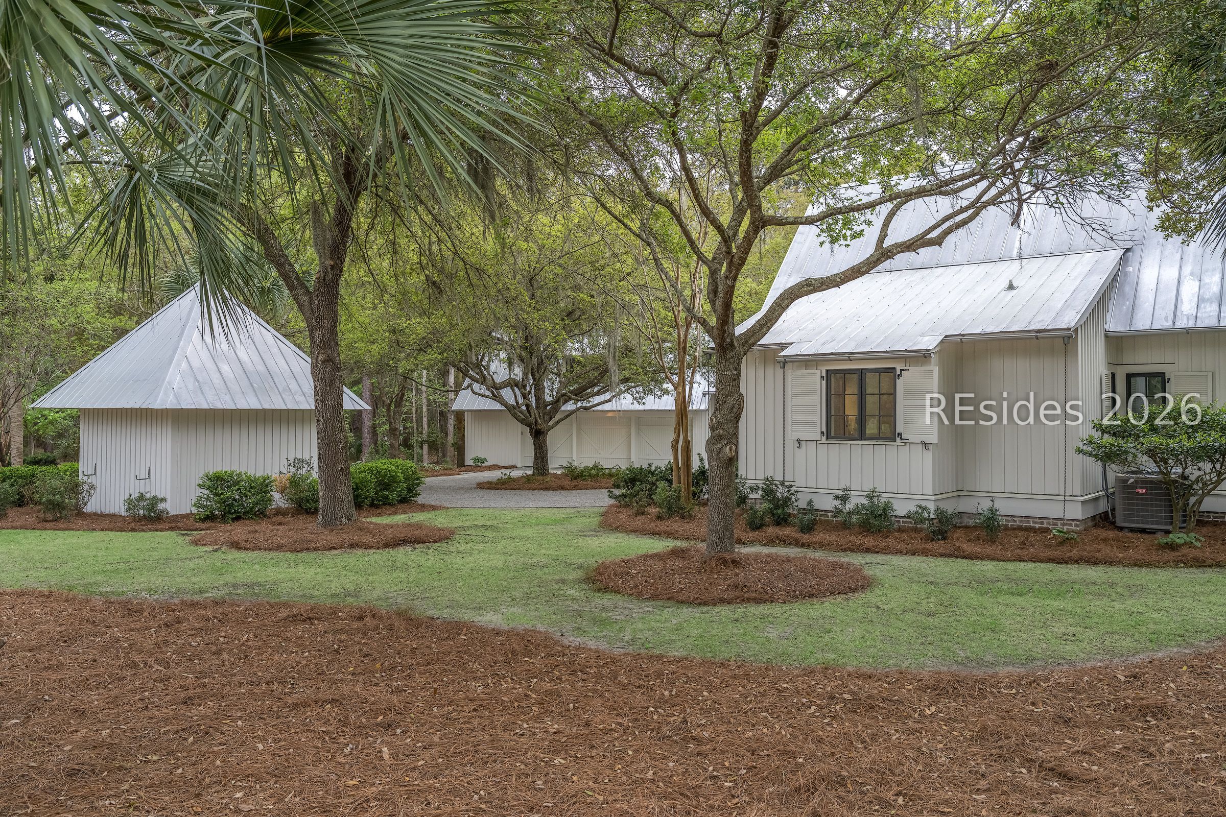 White metal-roofed home and detached outbuildings nestled among mature trees and landscaped grounds.