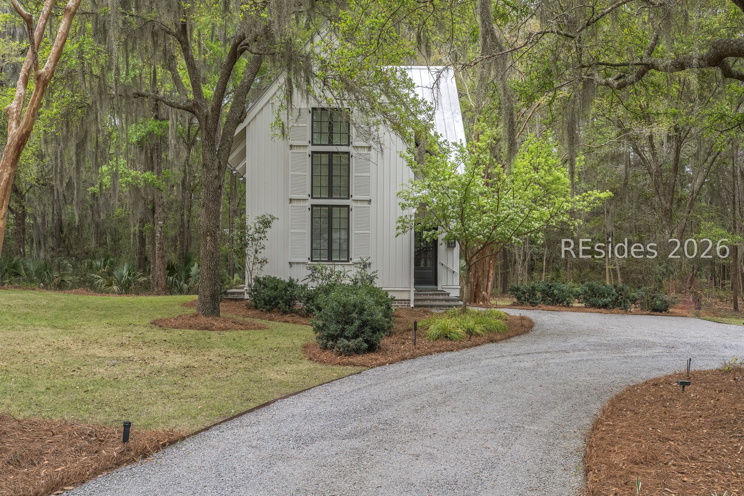 White house with dark-framed windows and a curved gravel driveway among moss-draped trees.