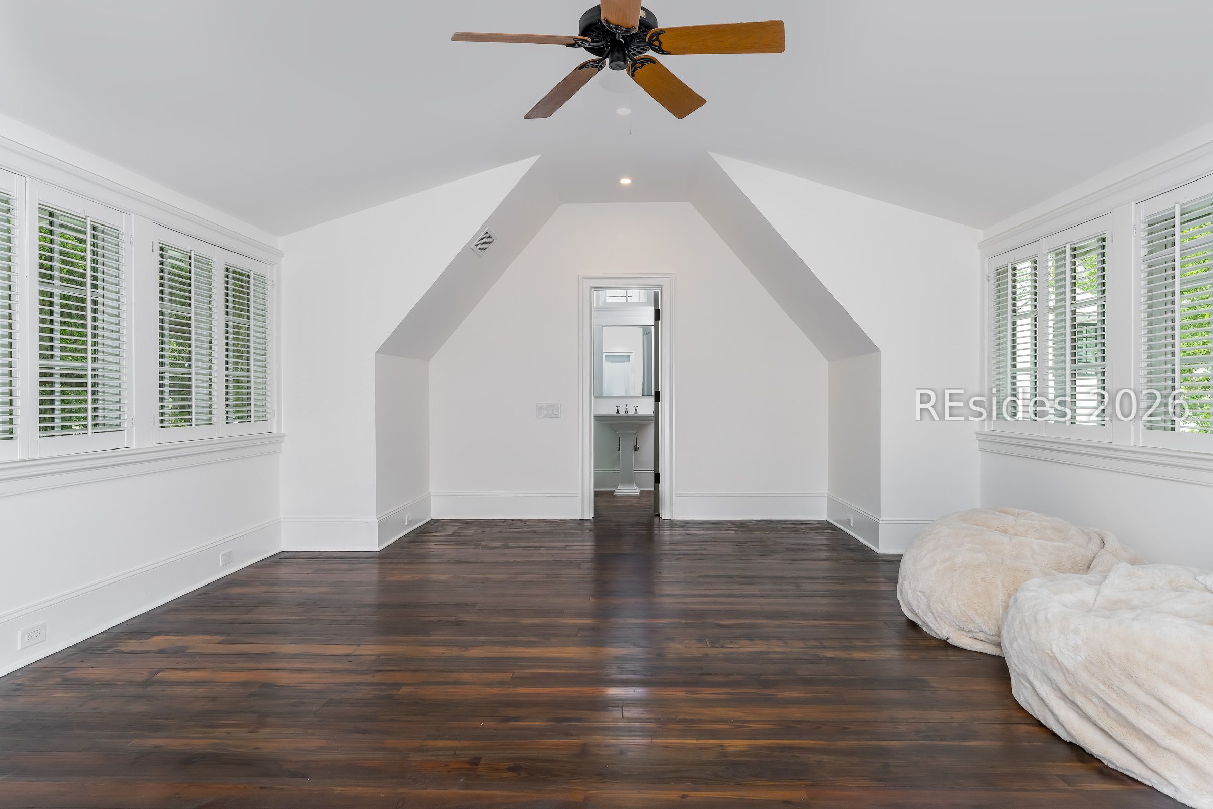Mostly empty bedroom with vaulted ceiling, dark wood floors, shuttered windows, and plush floor cushions.