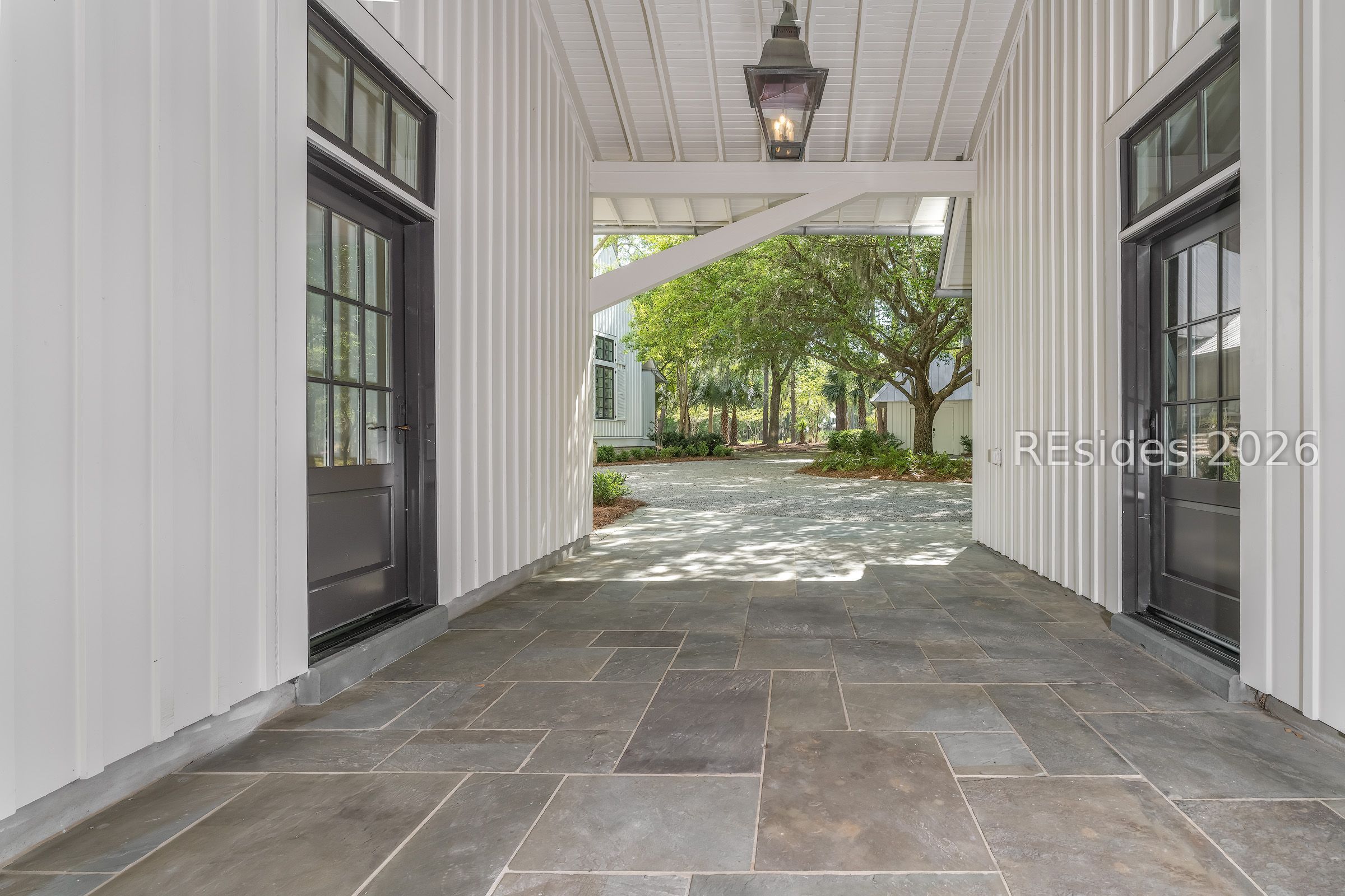 Covered entry porch with stone tile flooring, dark doors, and a tree-lined driveway beyond.
